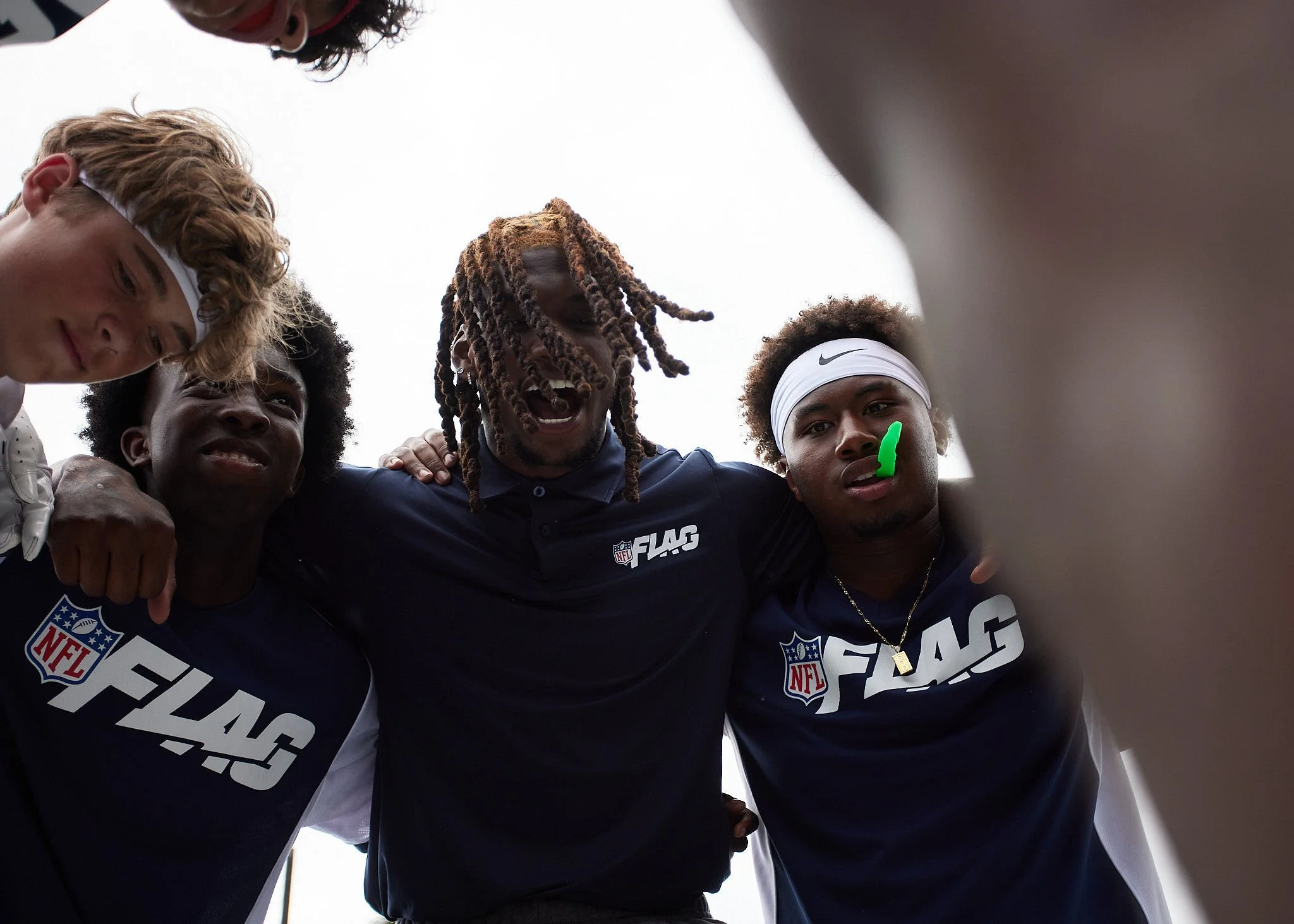A group of football players huddled together, seen from below, wearing NFL FLAG shirts, with one player in the center smiling and others looking focused.