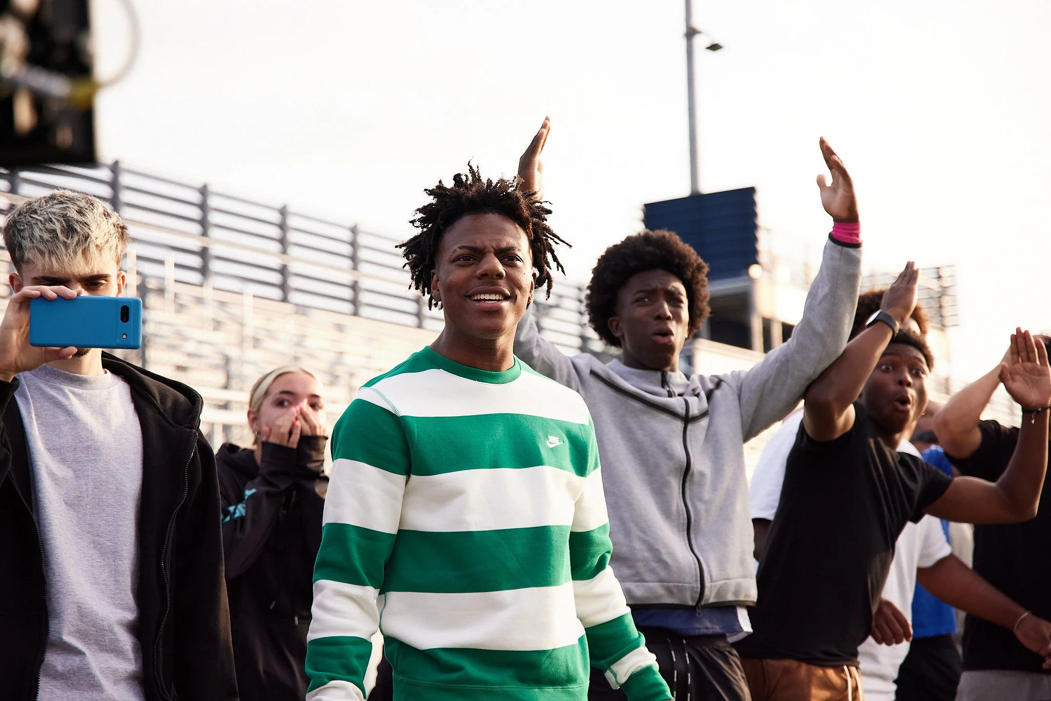 Group of diverse young people on a sports field, some raising their hands, one person taking a photo, others reacting with surprise or enthusiasm.