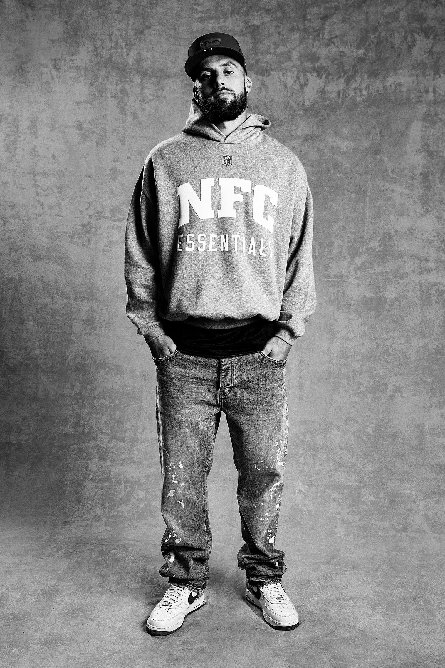 A man wearing a cap, hoodie with 'NFC ESSENTIALS' written on it, ripped jeans, and sneakers, standing against a textured wall.