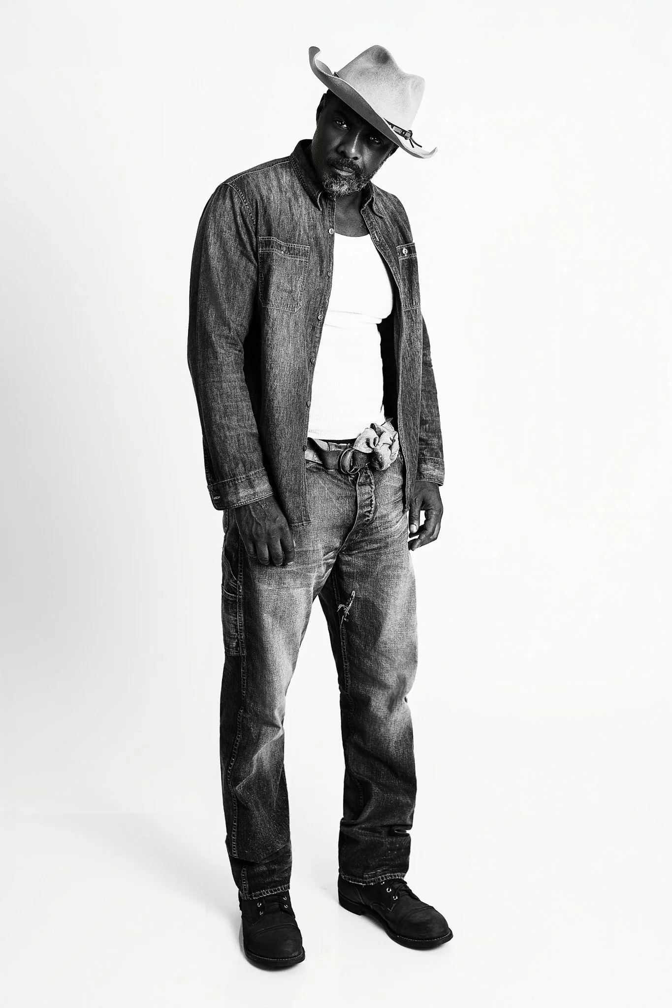 Black and white photo of a man wearing a cowboy hat, denim jacket, white shirt, jeans, and boots, standing against a plain background.