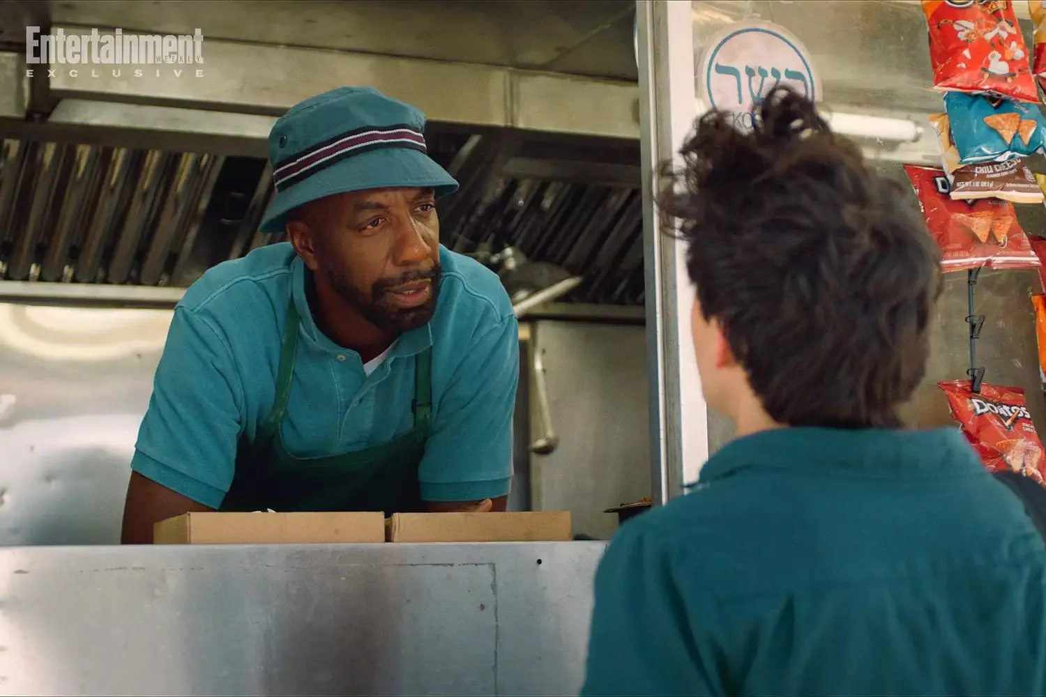 A man with a beard wearing a teal hat and shirt, talking to a person with dark, curly hair, inside a food truck or small eatery. Snacks like Doritos are visible on the side.