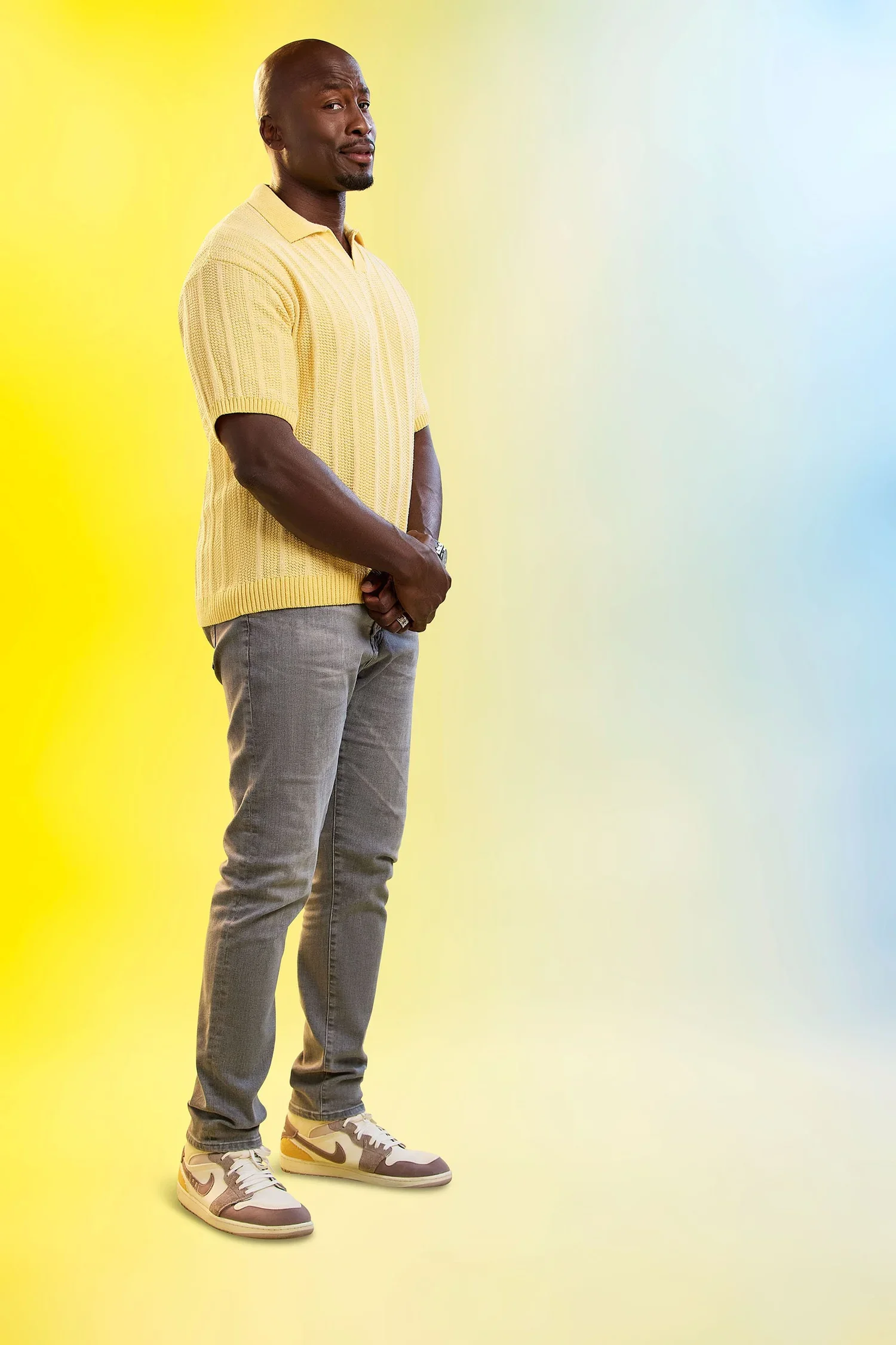 A man stands with a serious expression against a yellow and blue gradient background, wearing a yellow sweater, gray pants, and white sneakers.