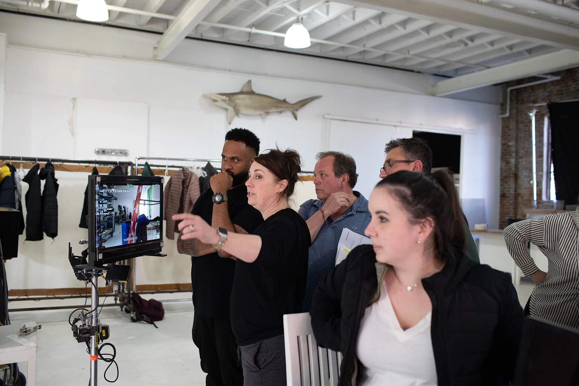 Group of five people in a studio setting, watching a monitor, with coats hanging in the background and a mounted fish on the wall.