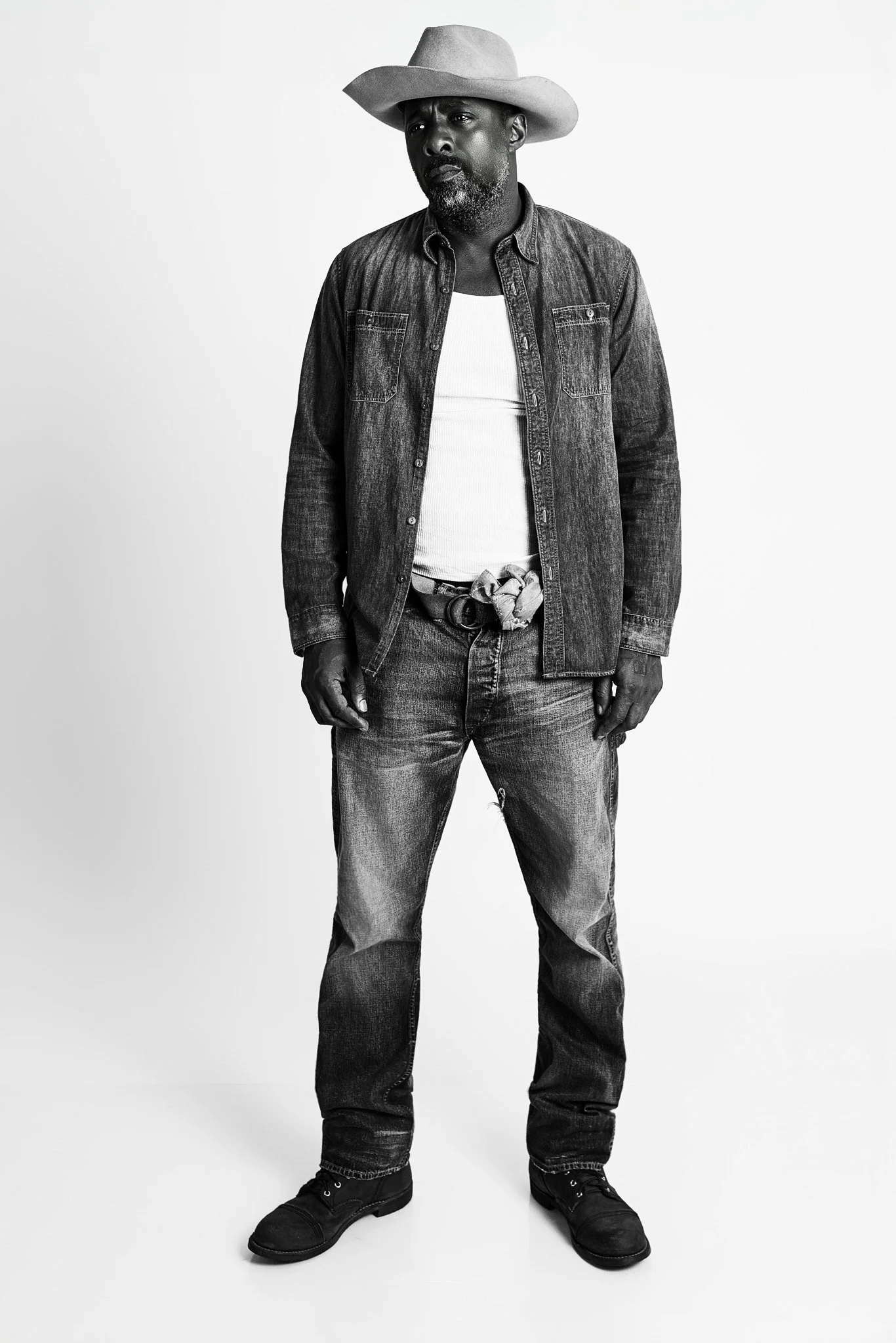 Black and white photo of a man wearing a wide-brimmed hat, denim shirt, white tank top, jeans, and boots, standing against a plain background.
