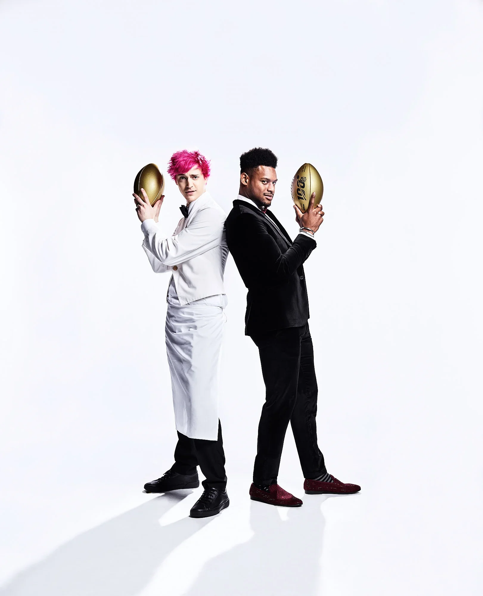 Two men in formal tuxedos stand back to back, each holding a golden rugby ball, against a plain white background.