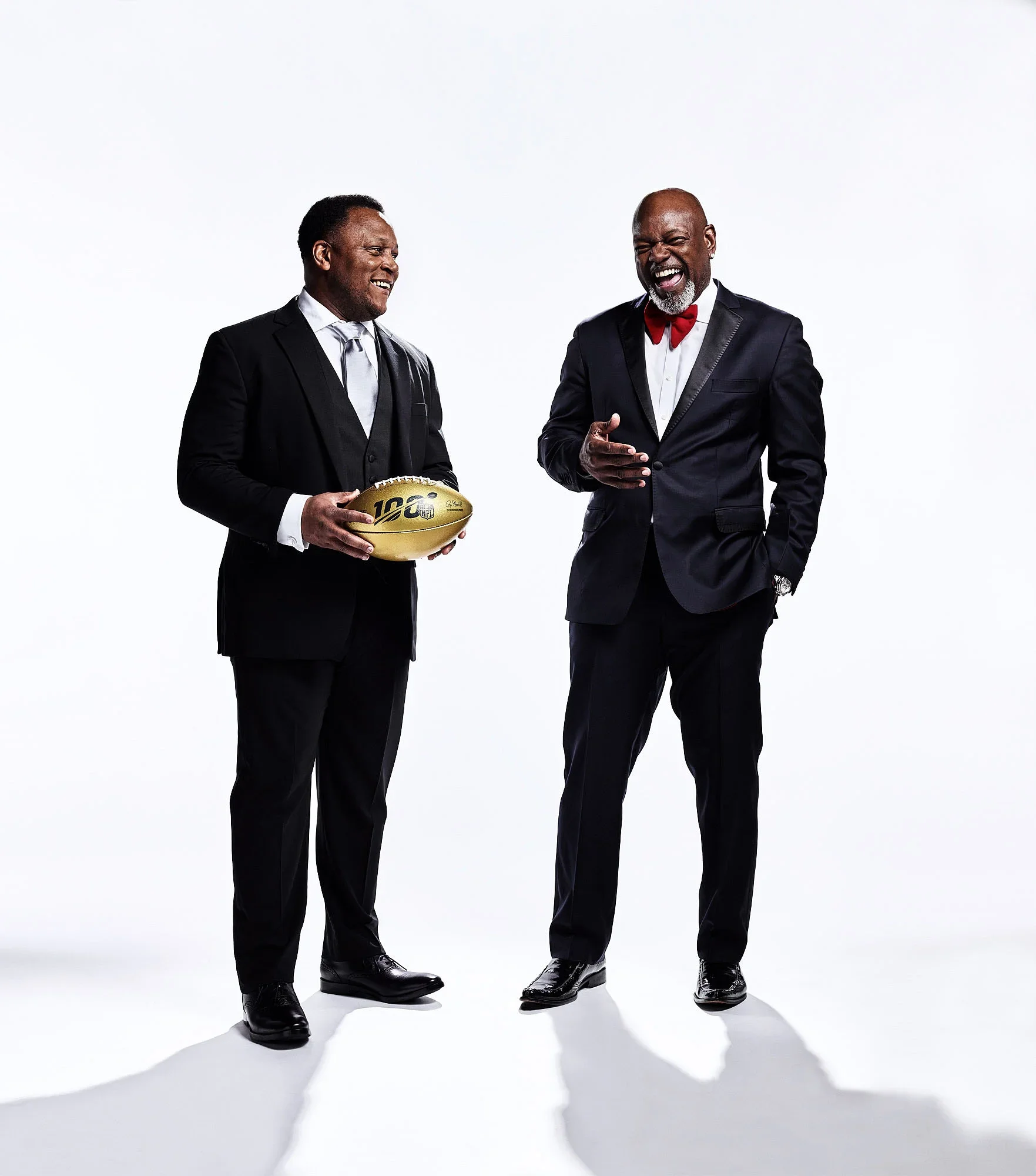 Two men in suits laughing and talking; one holding a gold football with '100' on it, against a plain white background.