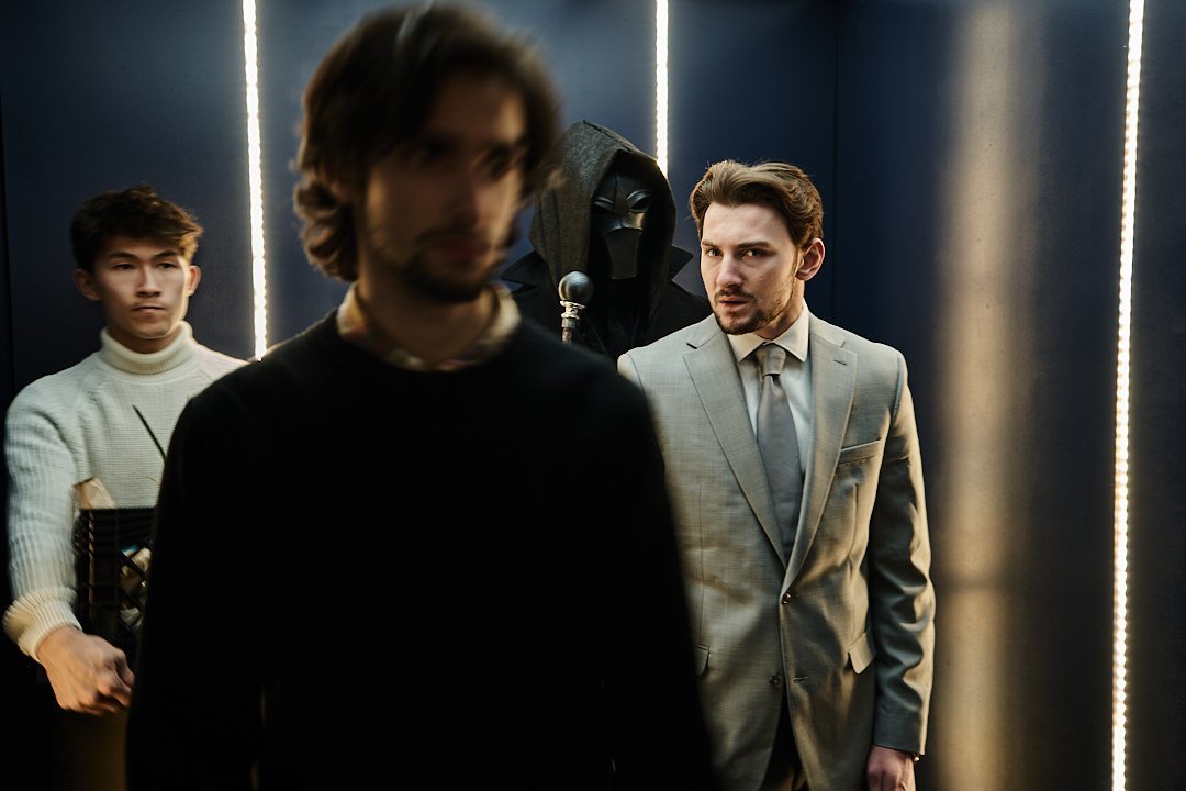 Four men in an elevator, with one person wearing a Darth Vader mask in the background. The man in the foreground is blurred, while the man in a gray suit looks puzzled. A young man in a white turtleneck stands to the left.