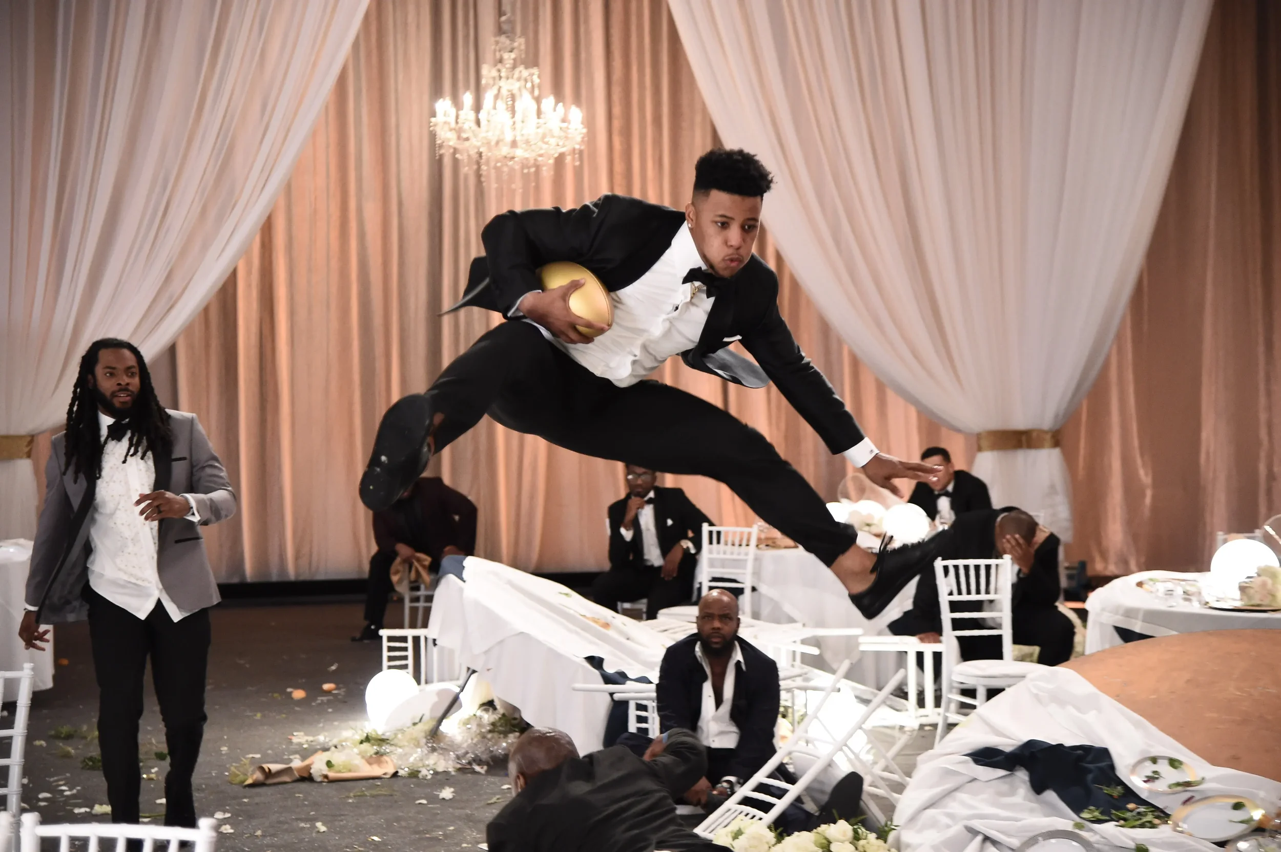 A man in a tuxedo is leaping over a banquet table at a formal event, holding a golden object, with people and broken chairs in the background.
