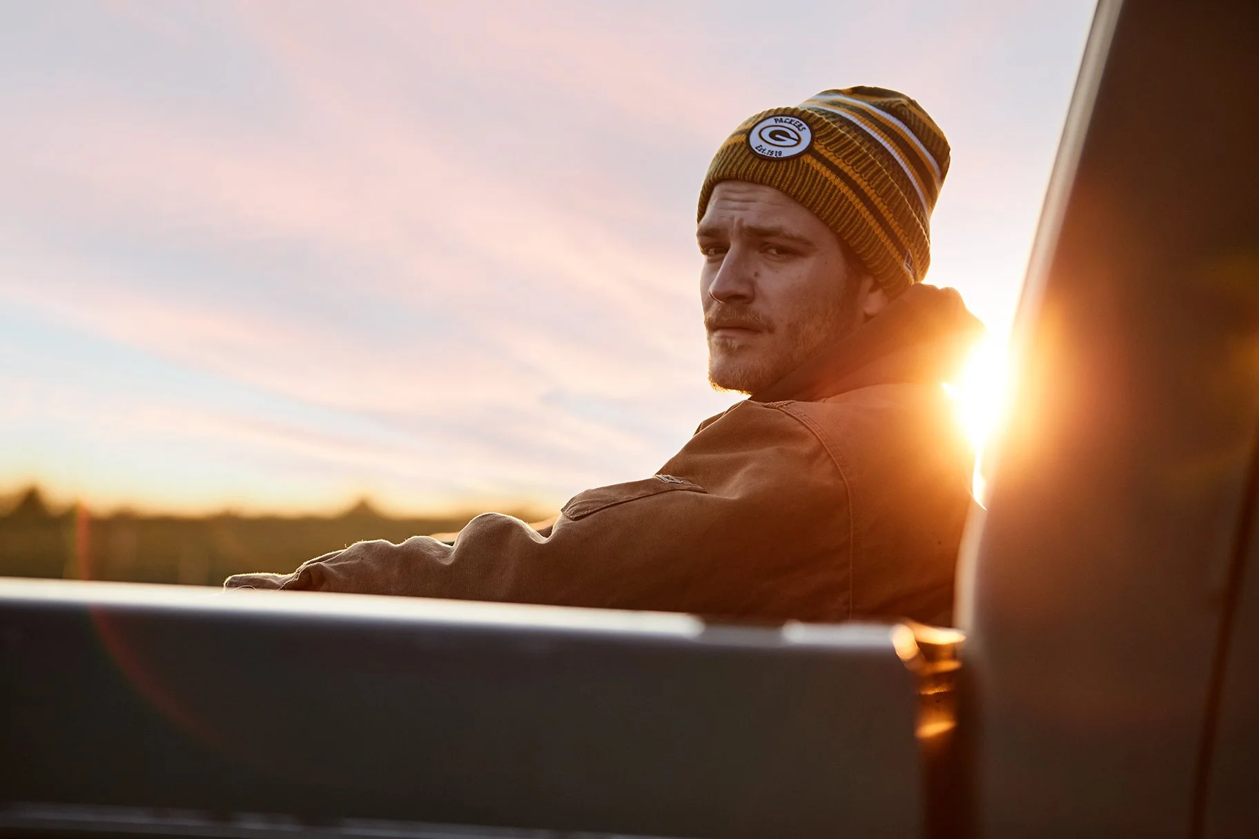 A man wearing a green Bay Packers beanie and a brown jacket sits by a car at sunset, looking over his shoulder.