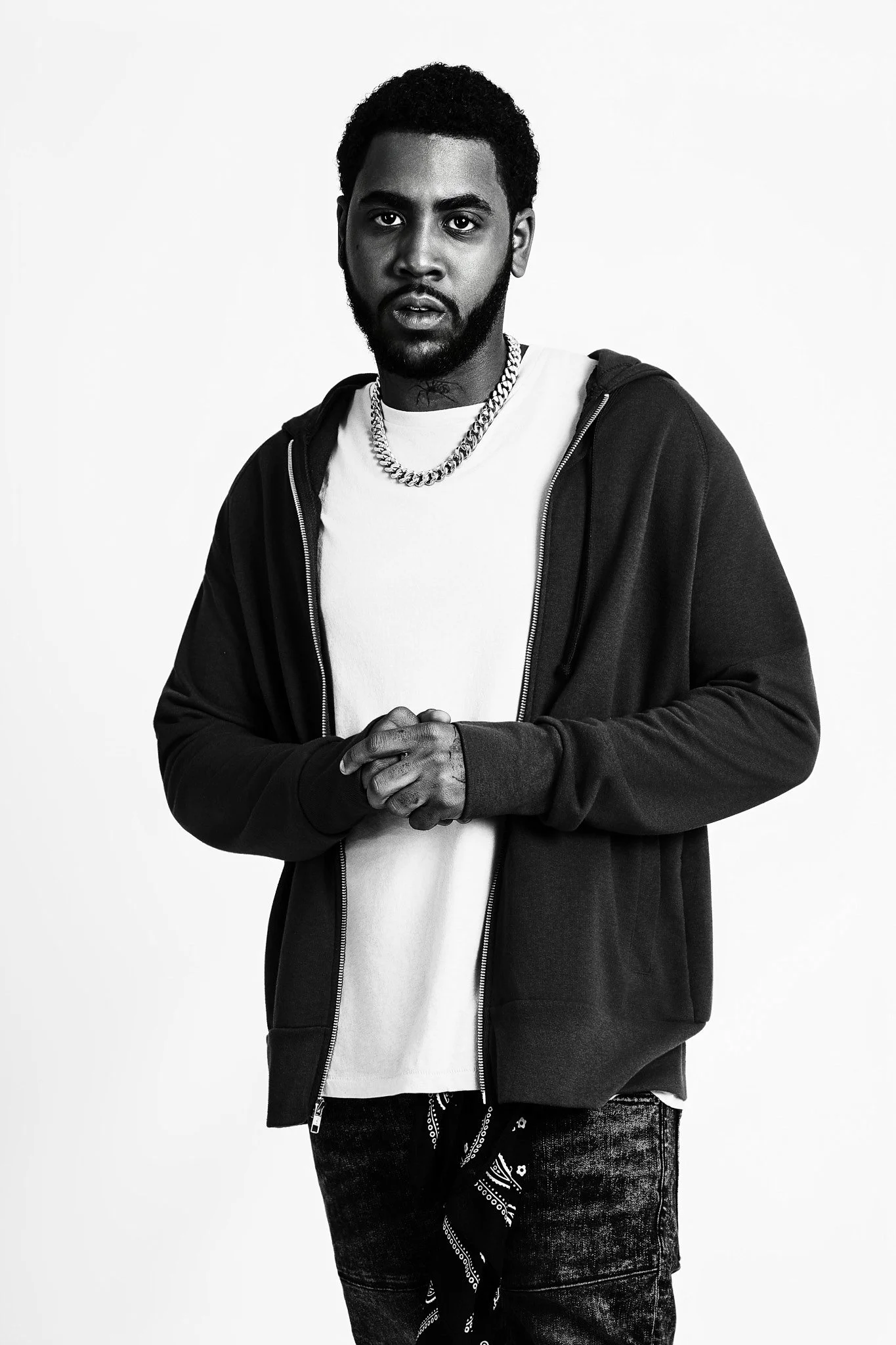 A young man with a beard and curly hair, wearing a white t-shirt, a black zip-up hoodie, a thick chain necklace, and jeans with a bandana hanging from his pocket, standing against a plain white background.