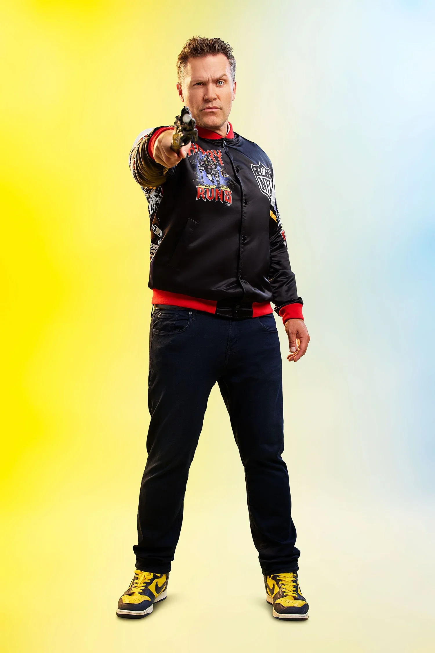 A man standing and pointing a gun directly at the camera, against a gradient background of yellow to blue.