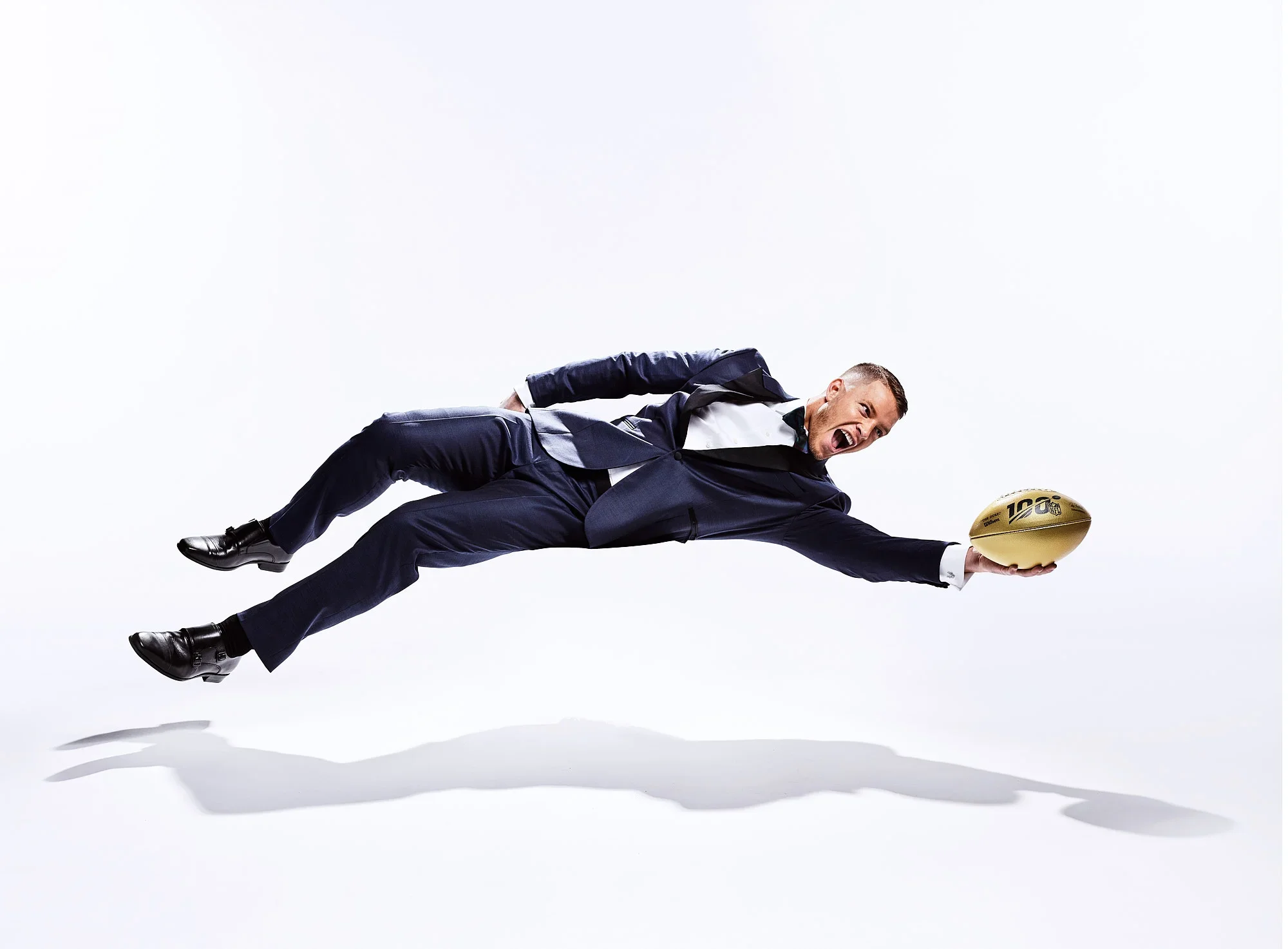 Man in a tuxedo lying horizontally and reaching out with a gold football in hand, with an excited expression