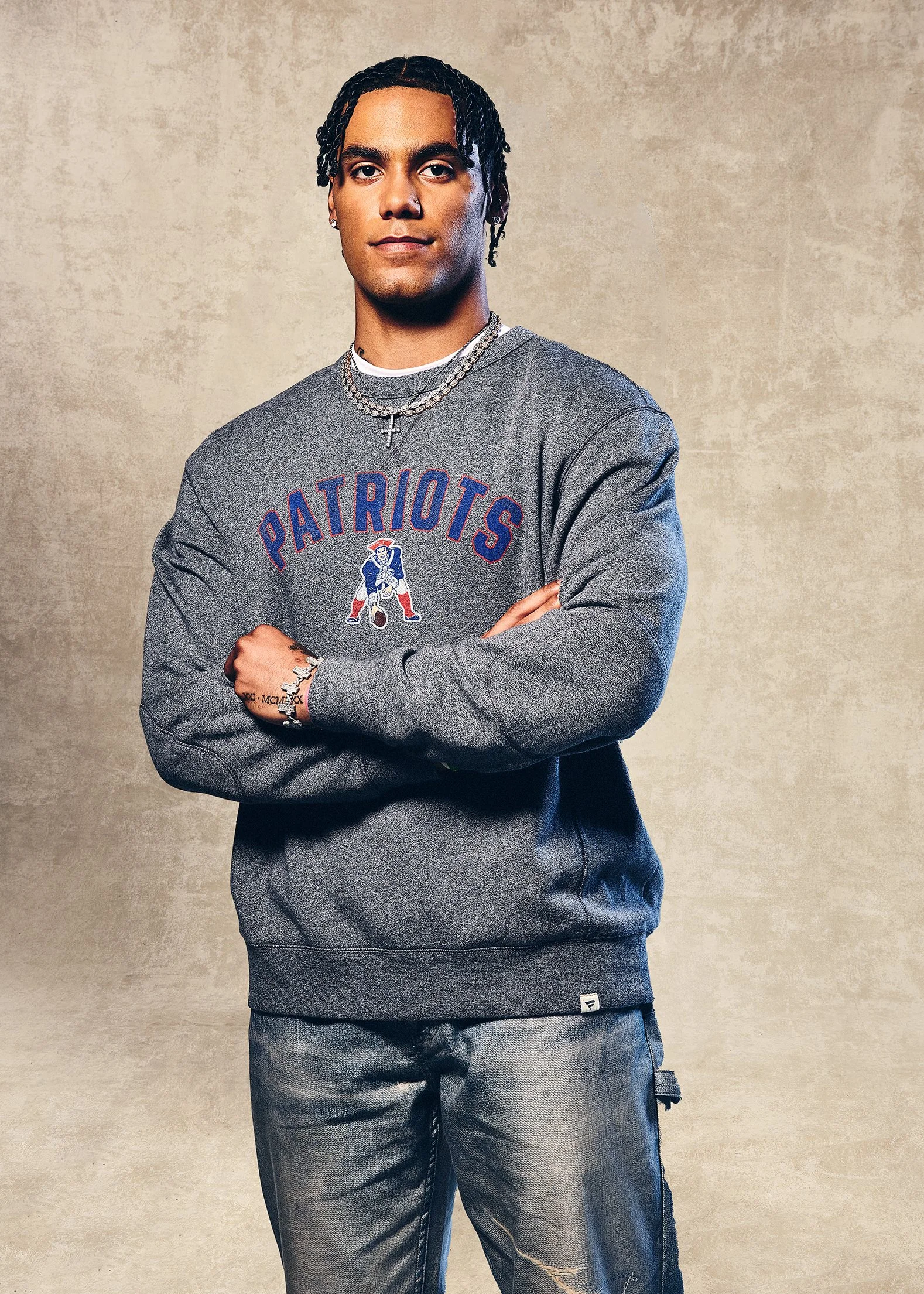 A young man with medium-length curly hair, wearing a gray Patriots sweatshirt, layered with necklaces, standing with arms crossed against a neutral background.