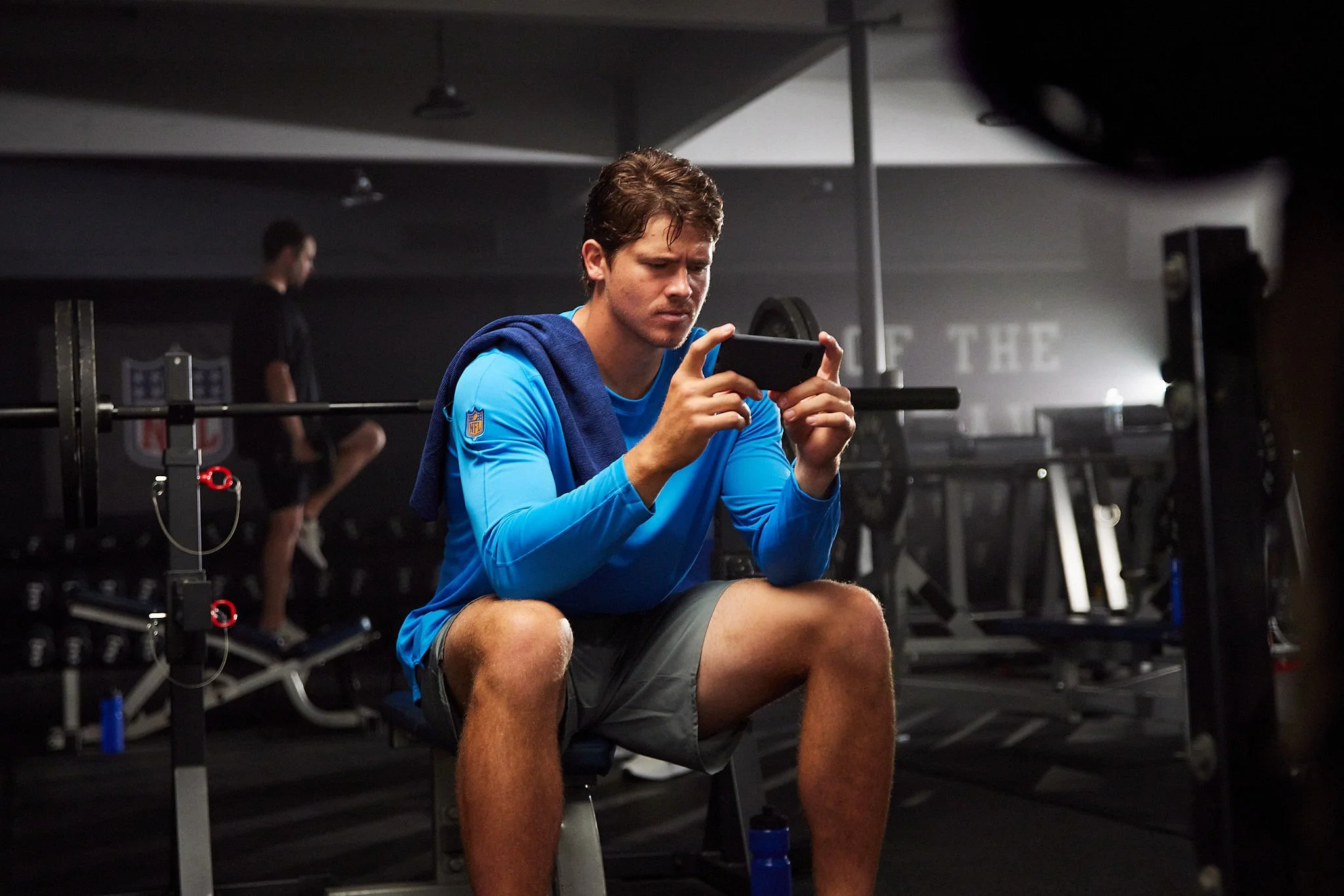 A young man in athletic clothing sits on a bench inside a fitness gym, looking at his phone. In the background, another person is sitting on gym equipment.