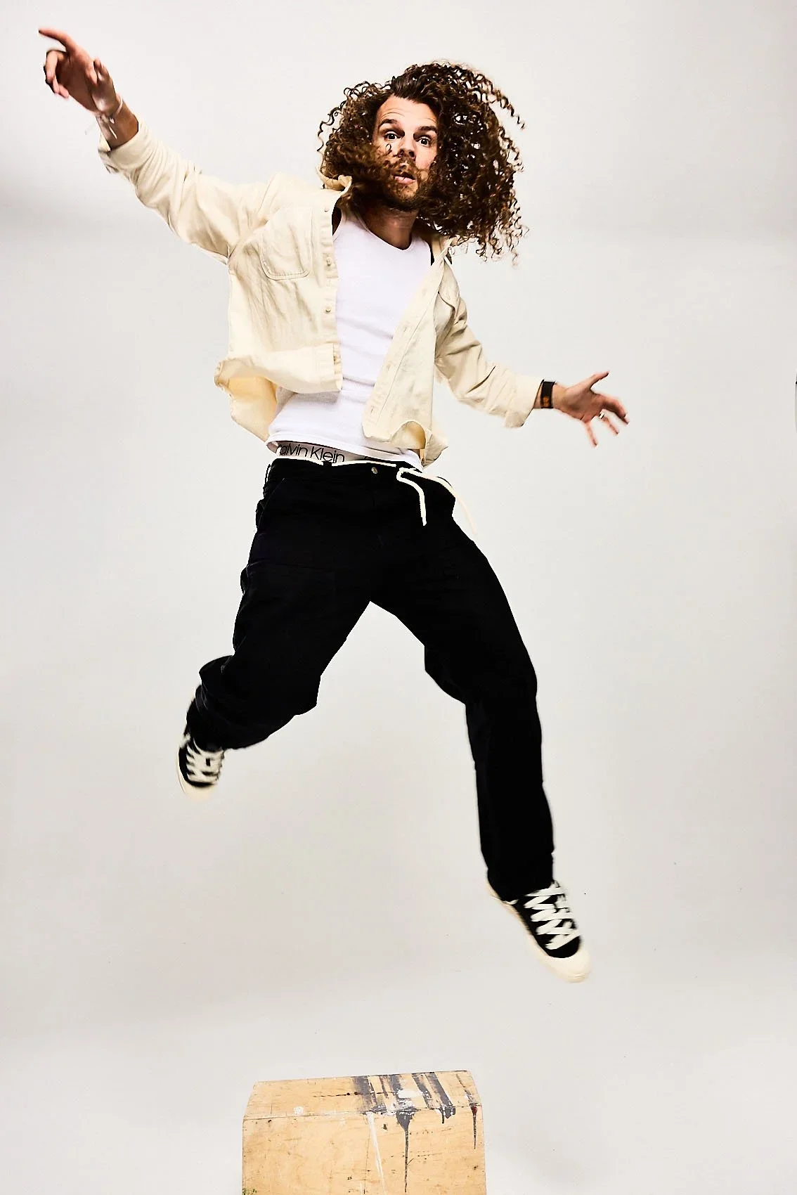A man with curly hair jumping in the air with a surprised expression, wearing a beige jacket, white shirt, black pants, and sneakers, against a plain background.