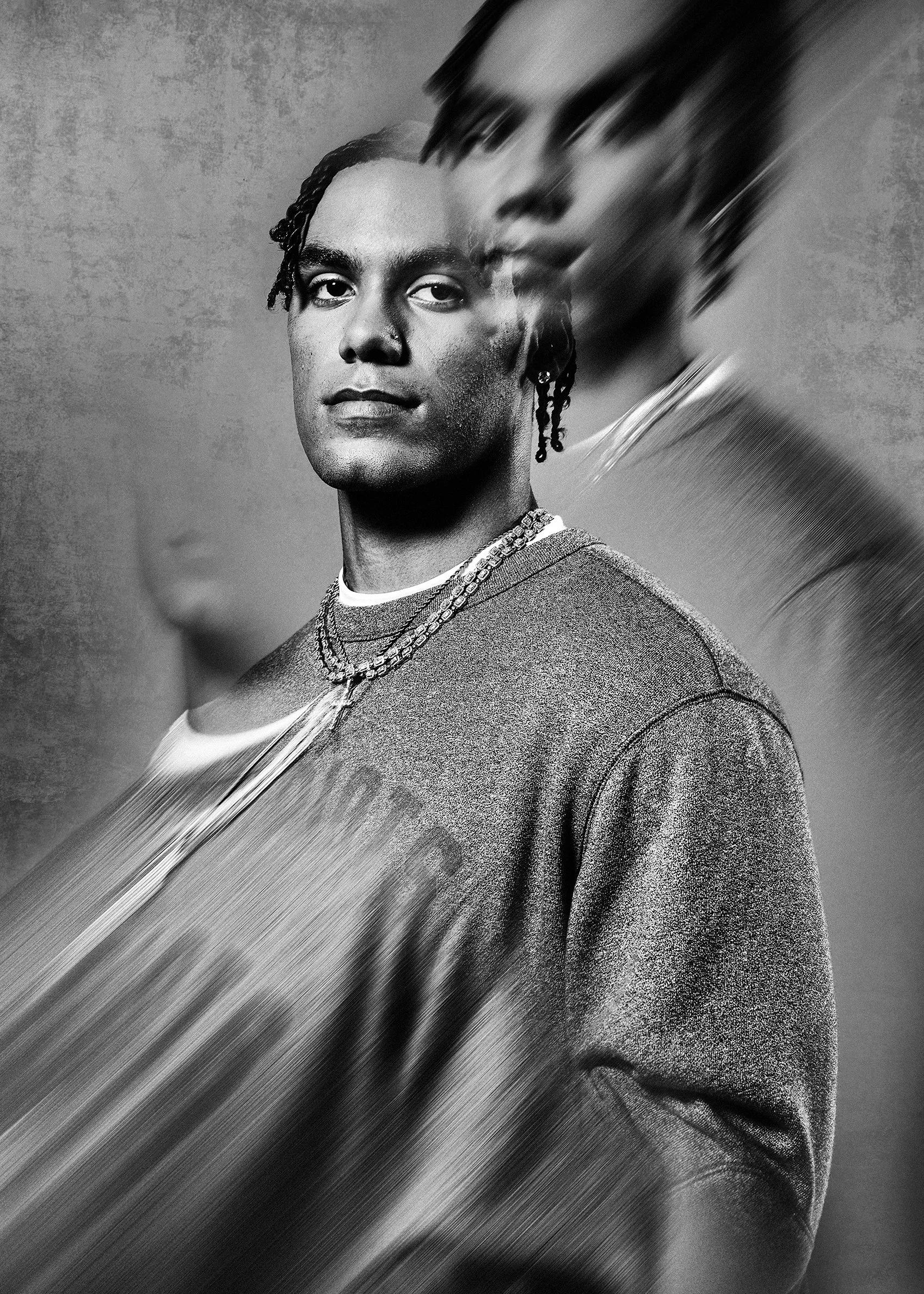 A black and white portrait of a young man wearing a sweater and layered necklaces, with a blurred and distorted background.