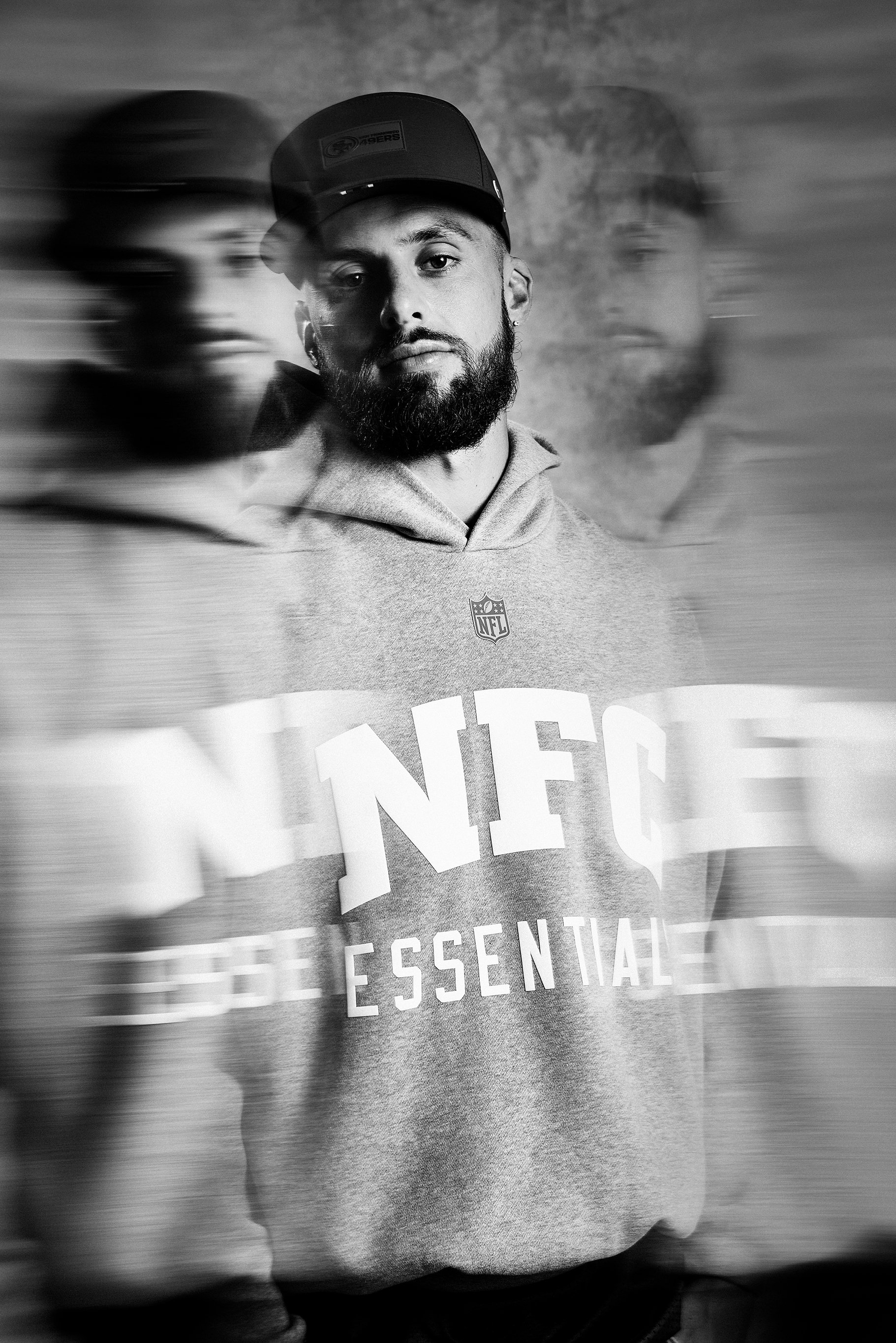 A black and white photo of a man with a beard, wearing a cap and a hoodie, with two blurred versions of himself in the background due to motion blur.