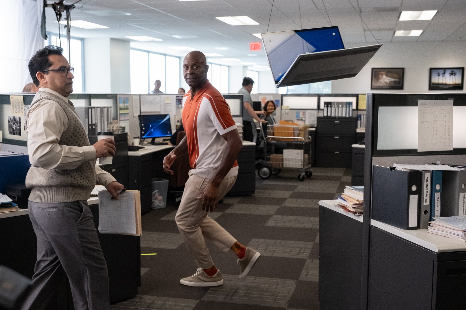Two men in an office interreacting, one holding a coffee cup and the other standing on one leg with hands by his side, with office cubicles, computers, and other employees in the background.