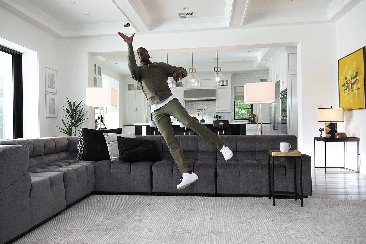 A person appears to be floating or jumping on a dark gray sectional sofa in a modern living room with an open kitchen in the background. The room has white walls, large windows, and contemporary decor.