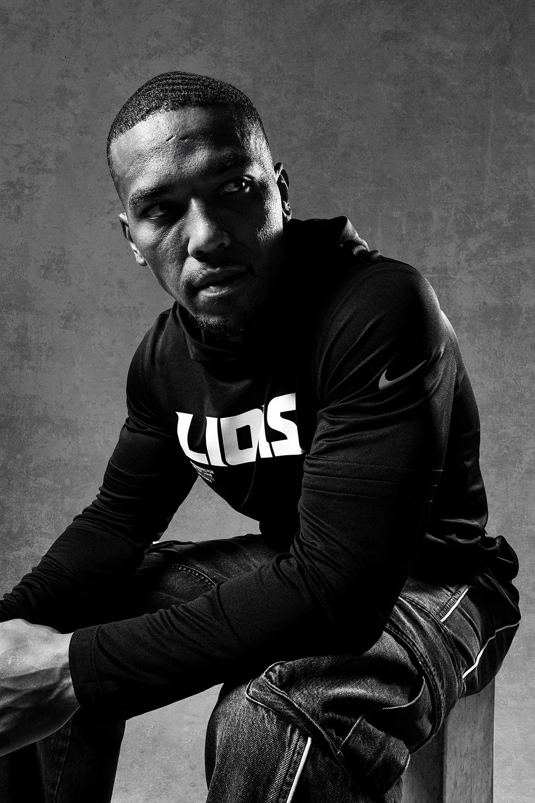 Black and white photo of a young man in casual clothing sitting against a textured background, looking to the side.