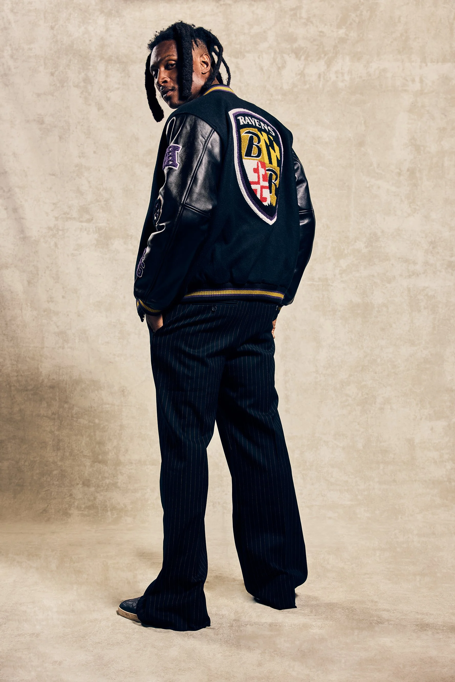 A man with dreadlocks wearing a Ravens varsity jacket, pinstripe pants, standing against a beige textured background.