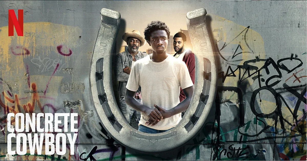 A group of four young men pose in front of a graffiti-covered wall with a large horseshoe-shaped object. The man in the foreground wears a white t-shirt and has short, curly hair. The other three men stand behind him, one with a hat and a gray jacket