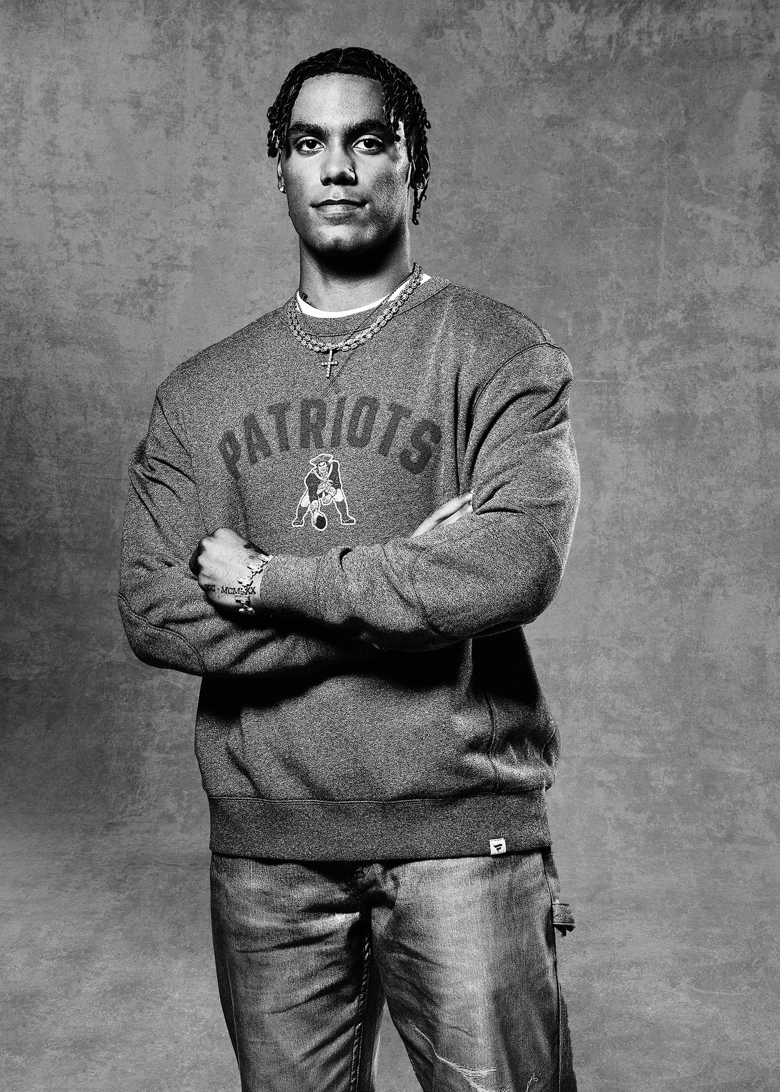 Black and white portrait of a young man with dreadlocks, wearing a sweatshirt with 'Patriots' and a logo, layered necklaces, and jeans, standing against a plain background.