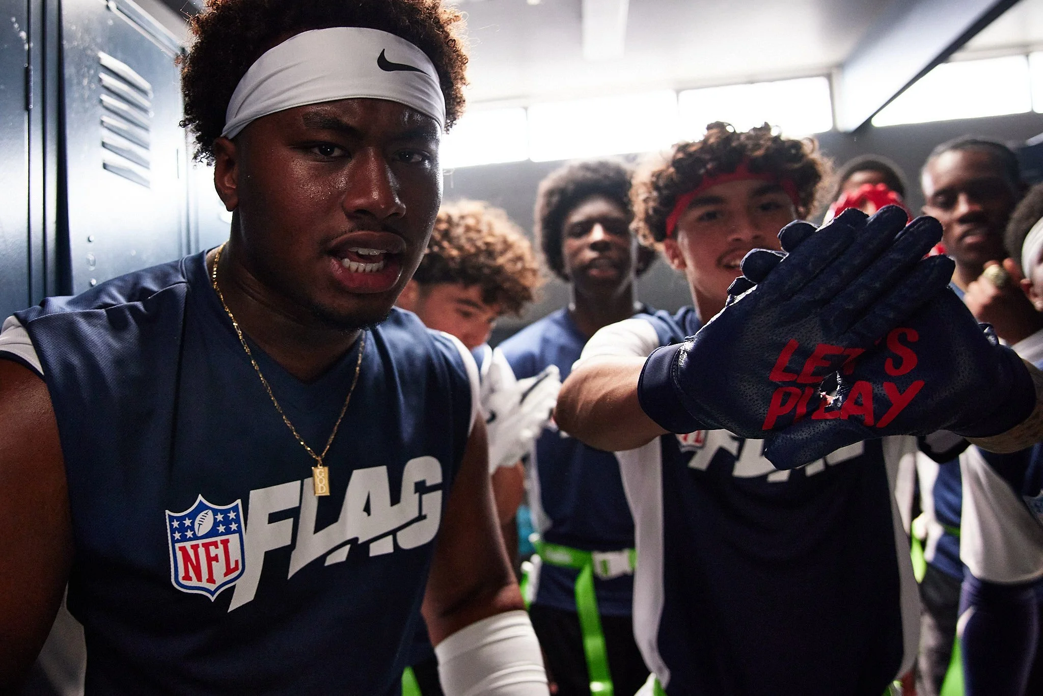 Group of football players in locker room, wearing NFL FLAG t-shirts, with one player in the foreground wearing a white headband and a gold necklace, raising his hand with gloves that say "LET'S PLAY".
