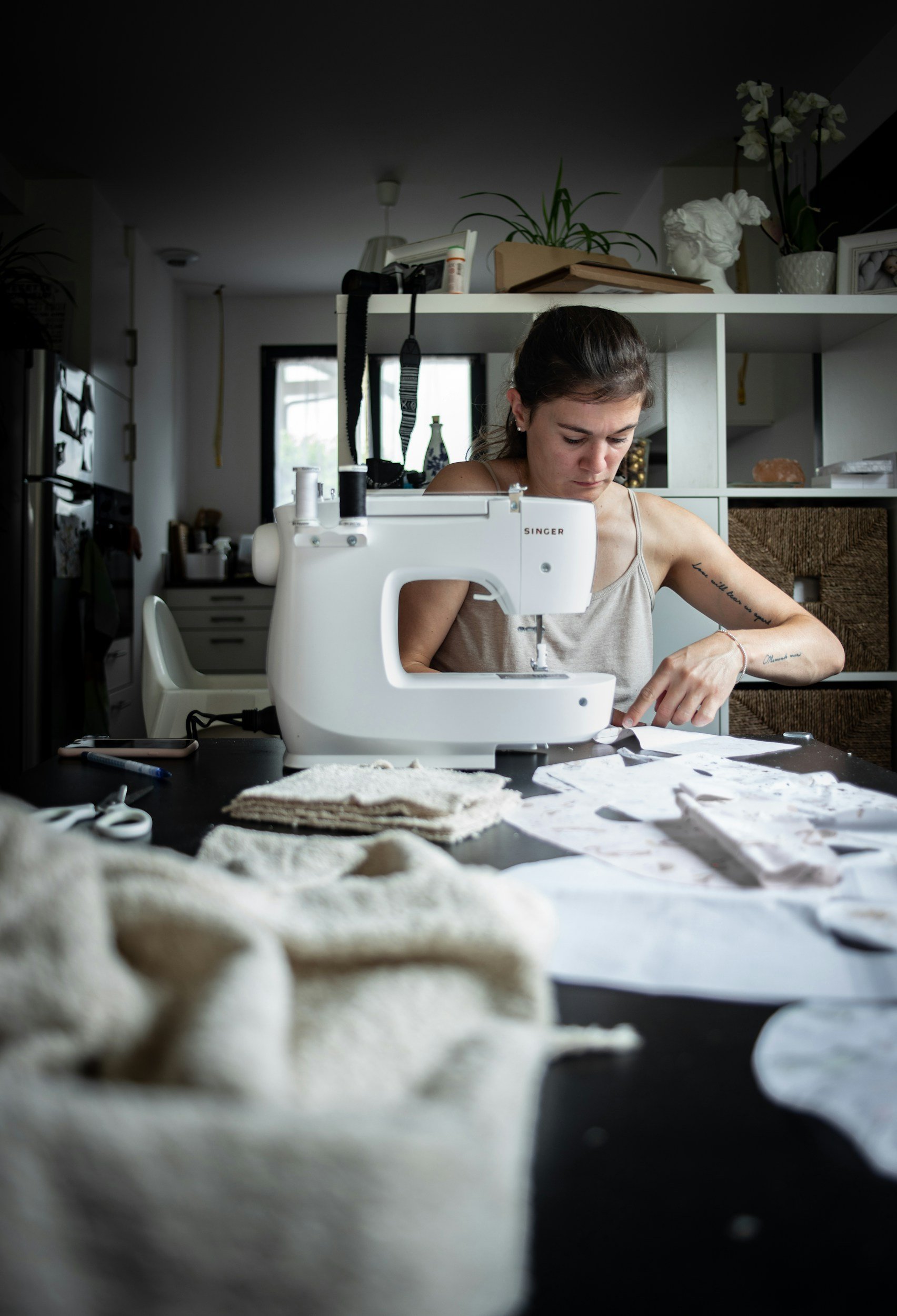 White person wearing tank top sitting in front of sewing machine. Fabric and paper scattered on table