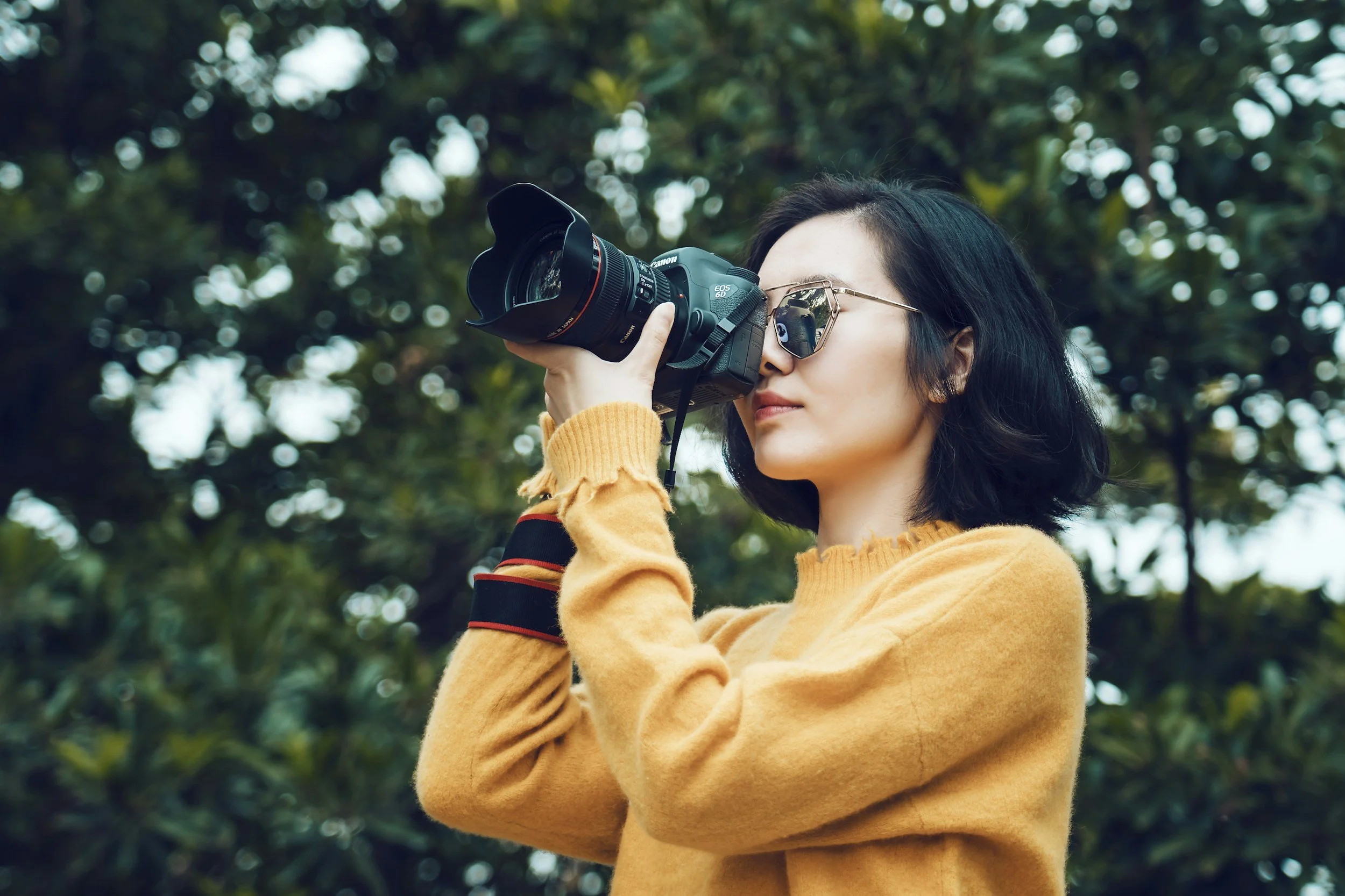 Asian woman with shoulder length black hair wearing sunglasses and yellow sweatshirt looking through professional camera. Trees in background