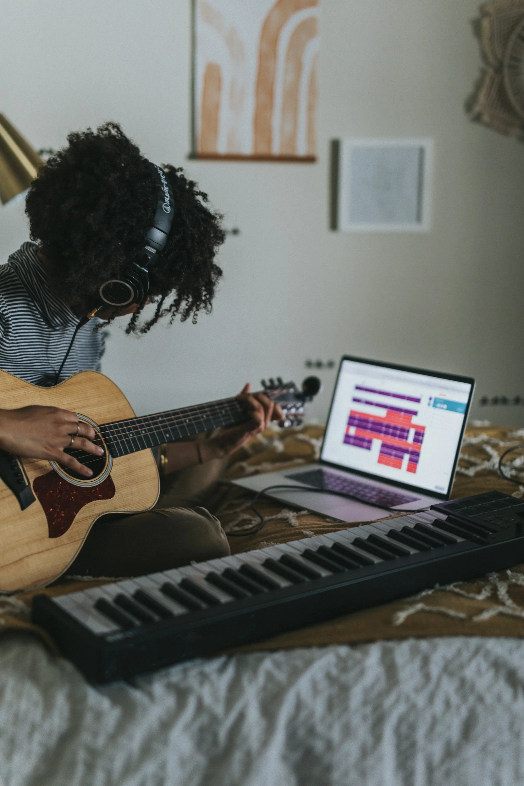 Black woman playing guitar and wearing headphones while sitting on bed. A laptop and keyboard lay in front of her