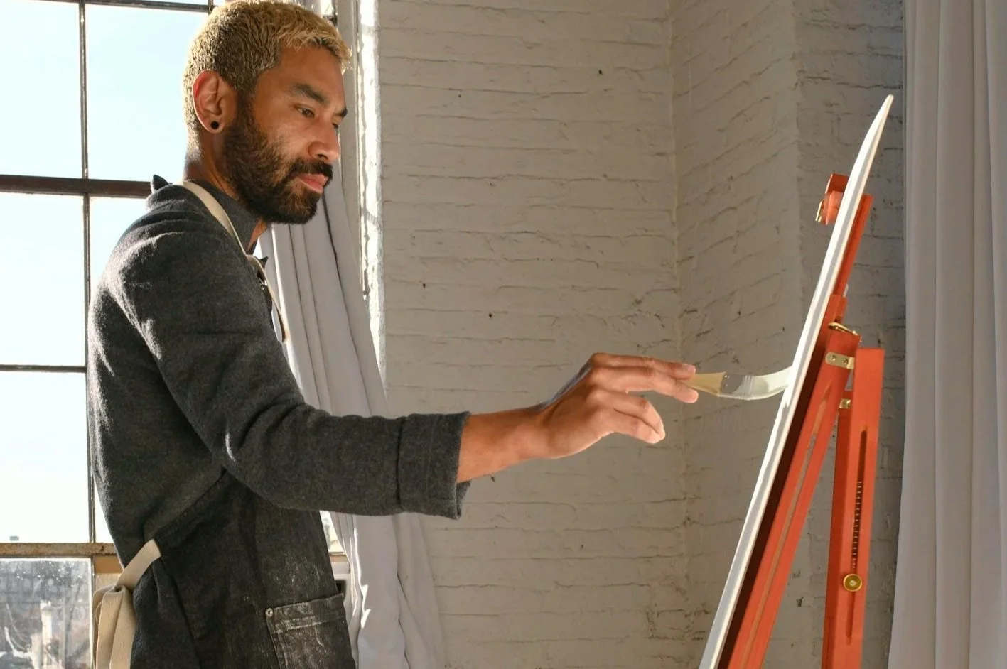 Side view of man with beard and brown skin painting at an easel in a sunny room