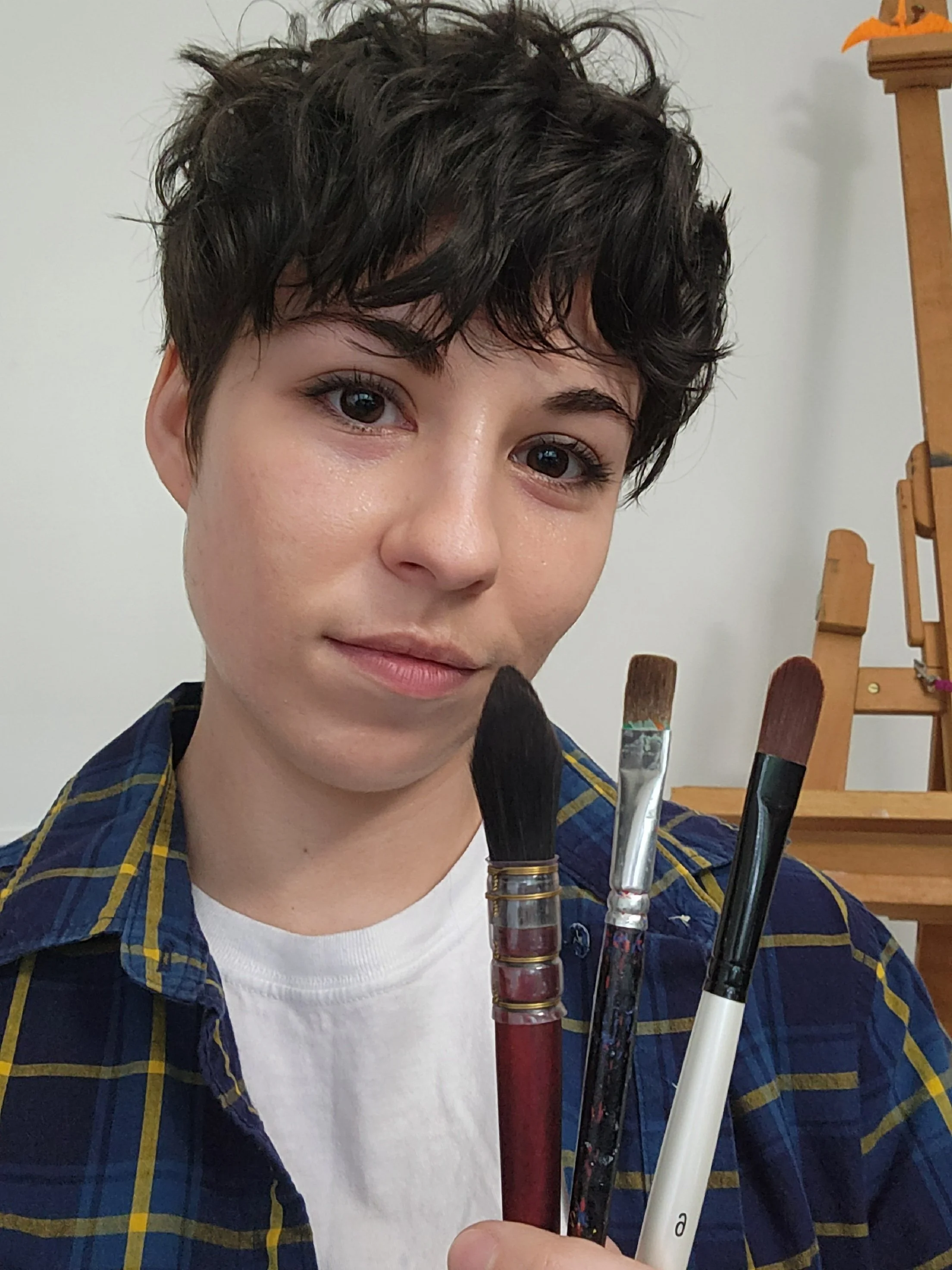 Headshot of woman with short black hair smiling wearing blue and yellow flannel holding three paint brushes. Easel in background