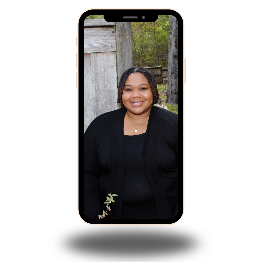 A smiling woman taking a selfie outdoors with trees and a wooden structure in the background.