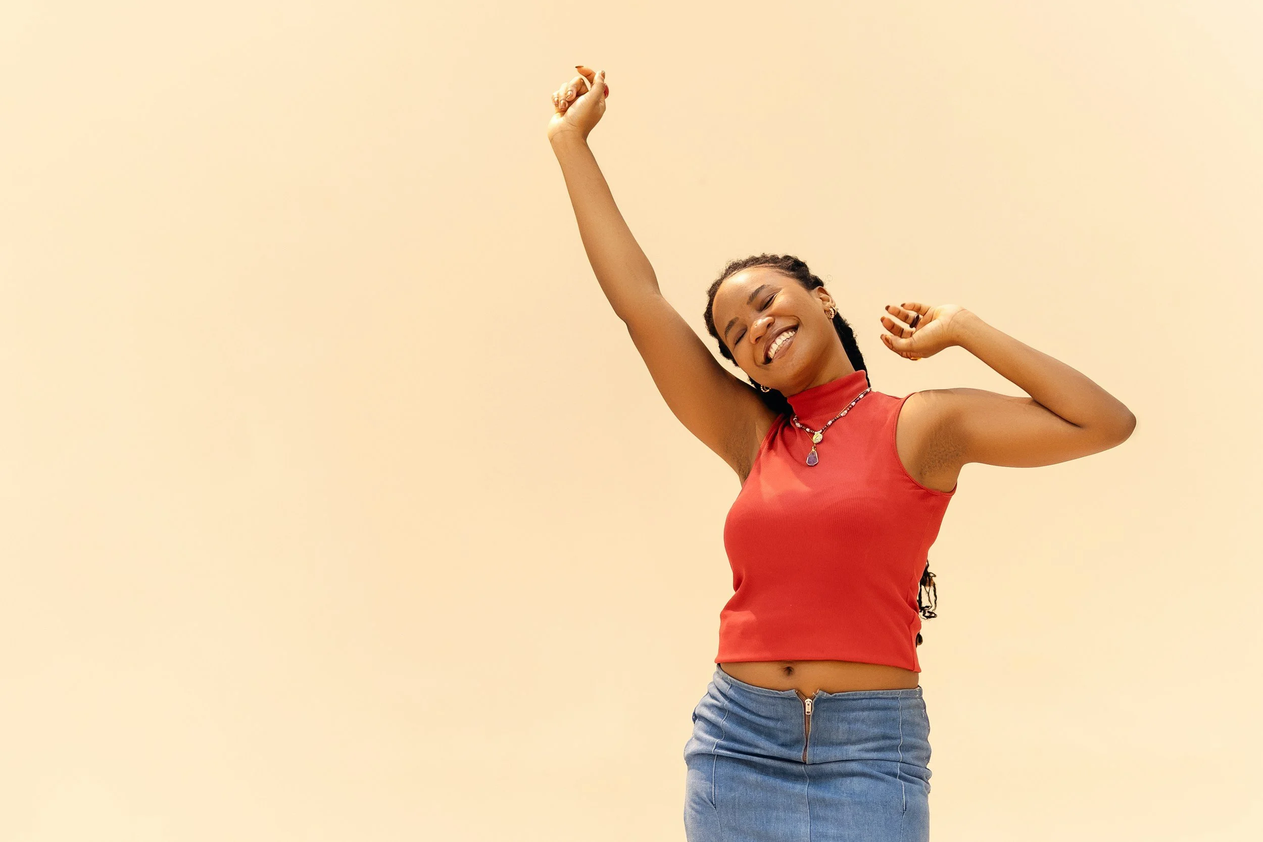 A young woman with braids is smiling and dancing with her eyes closed. She has an arm raised above her head and the other bent at her side. She is wearing a red sleeveless top, a jean skirt, and jewelry, including a necklace with a purple pendant.