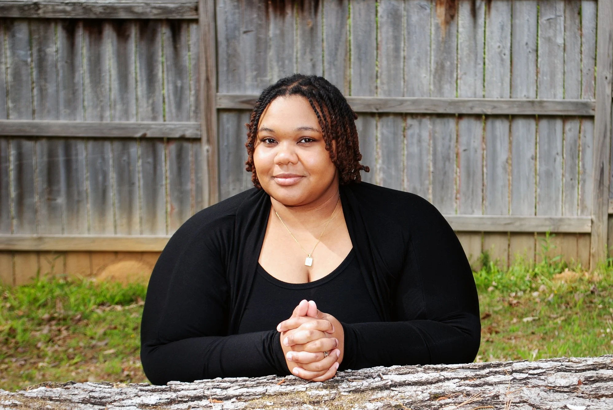 A woman with shoulder-length curly hair, wearing a black top and a gold necklace, sitting outdoors with a wooden fence and grass in the background.