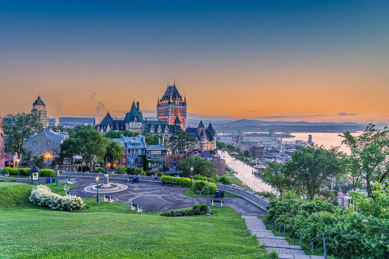 Vue panoramique de Québec avec le château Frontenac au coucher du soleil, bâtiments historiques, rivière et paysage verdoyant.