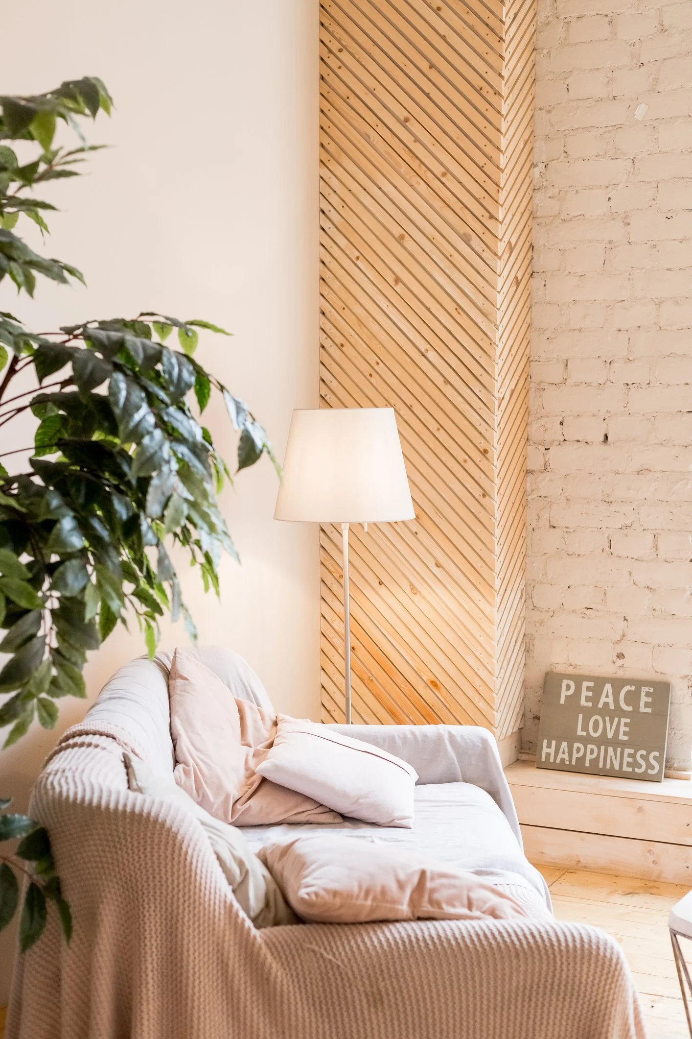 Un salon intérieur avec un canapé beige, des coussins, une lampe sur pied, un mur en briques blanches et un panneau en bois décoratif, avec une affiche qui dit « PEACE LOVE HAPPINESS ».