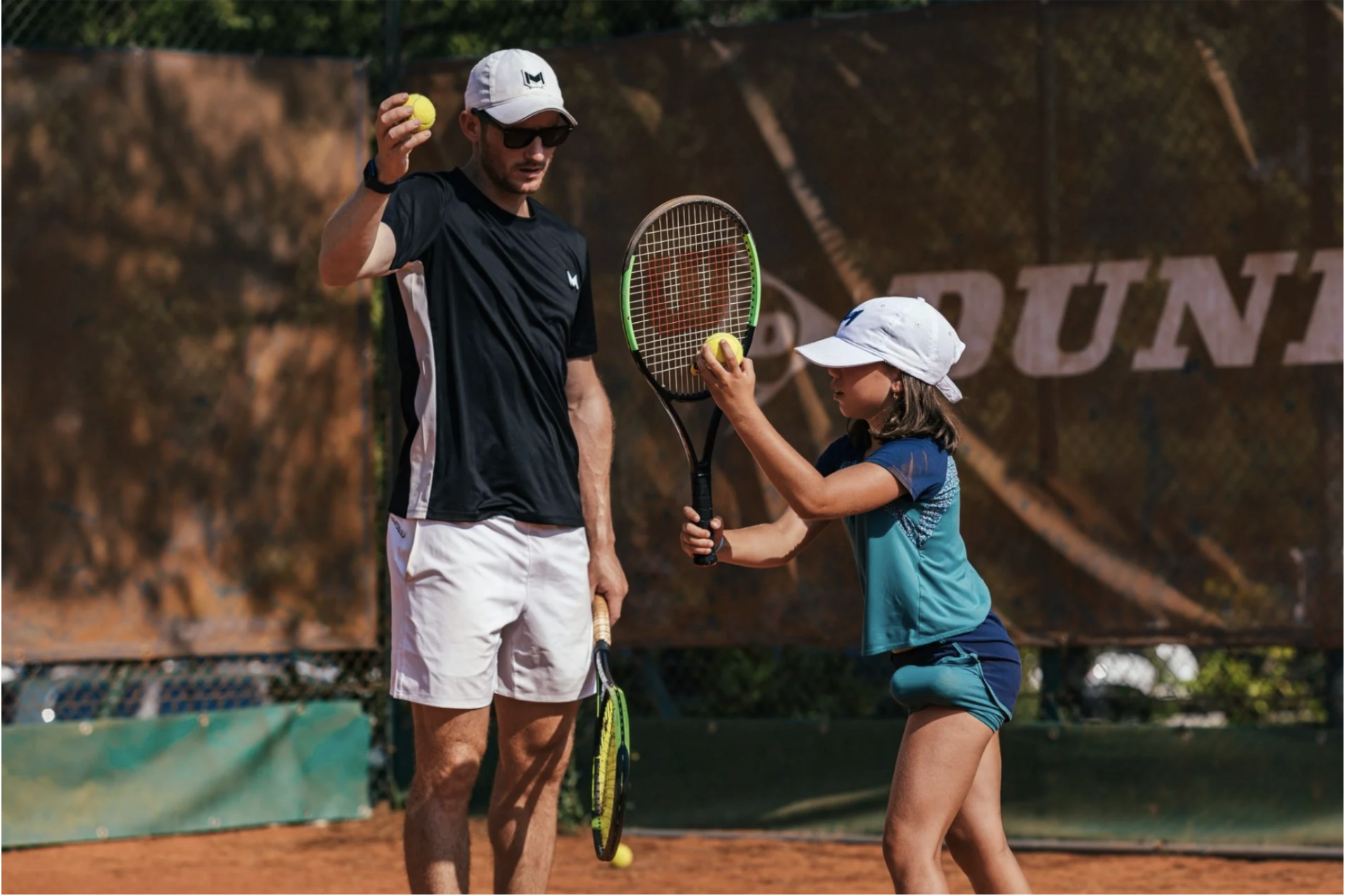 A man and a young girl on a tennis court, both holding tennis rackets and tennis balls. The man is wearing sunglasses, a white cap, a navy shirt, and white shorts. The girl is wearing a white cap, a blue and teal shirt, and teal shorts. They appear to be practicing or having a lesson outdoors.