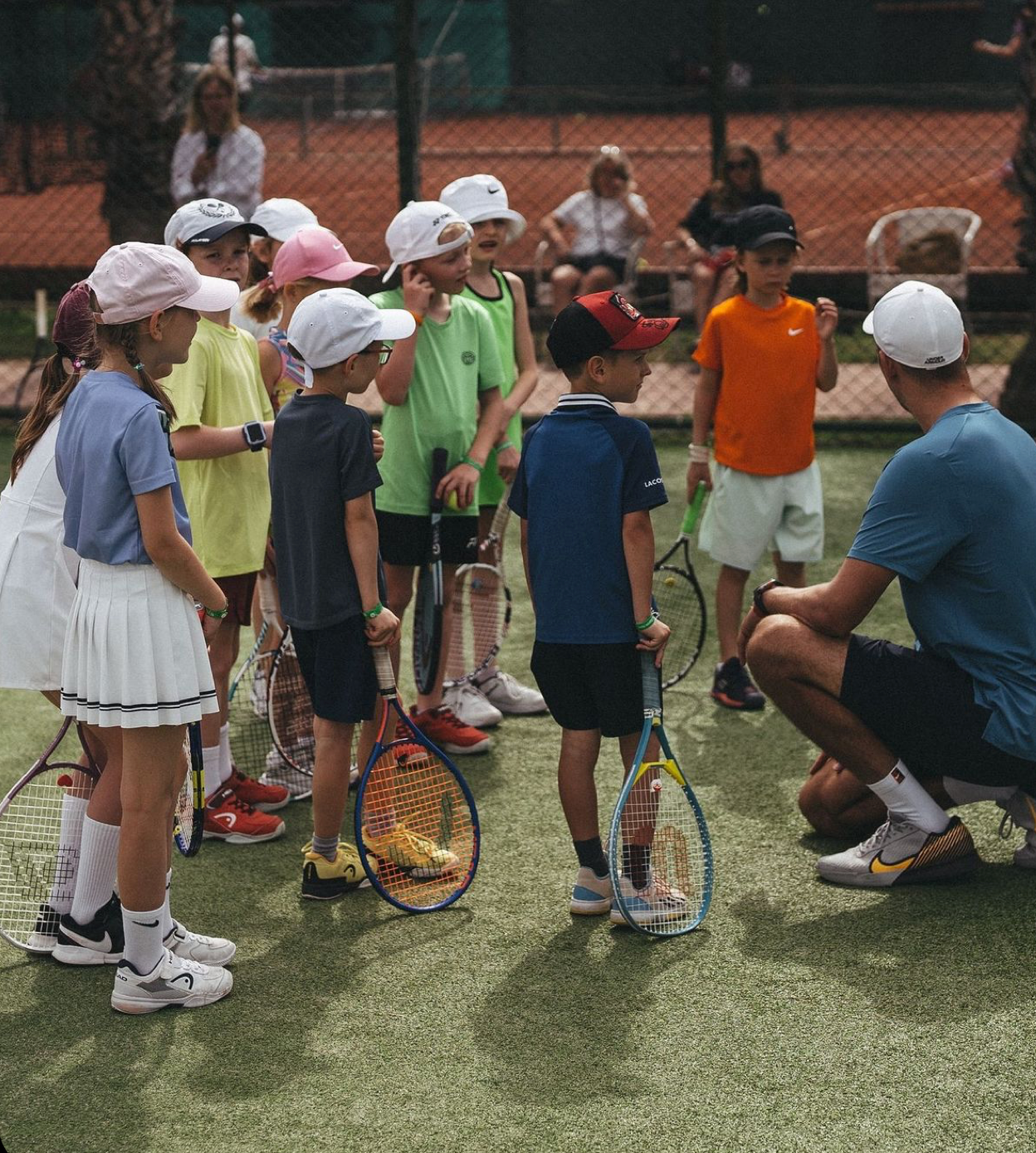 Group of children on a tennis court receiving instruction from an adult coach. The children are holding tennis rackets and wearing tennis caps.