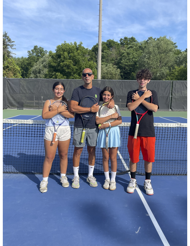 A coach and three young tennis players standing on a tennis court, holding tennis rackets, with a black net and trees in the background.