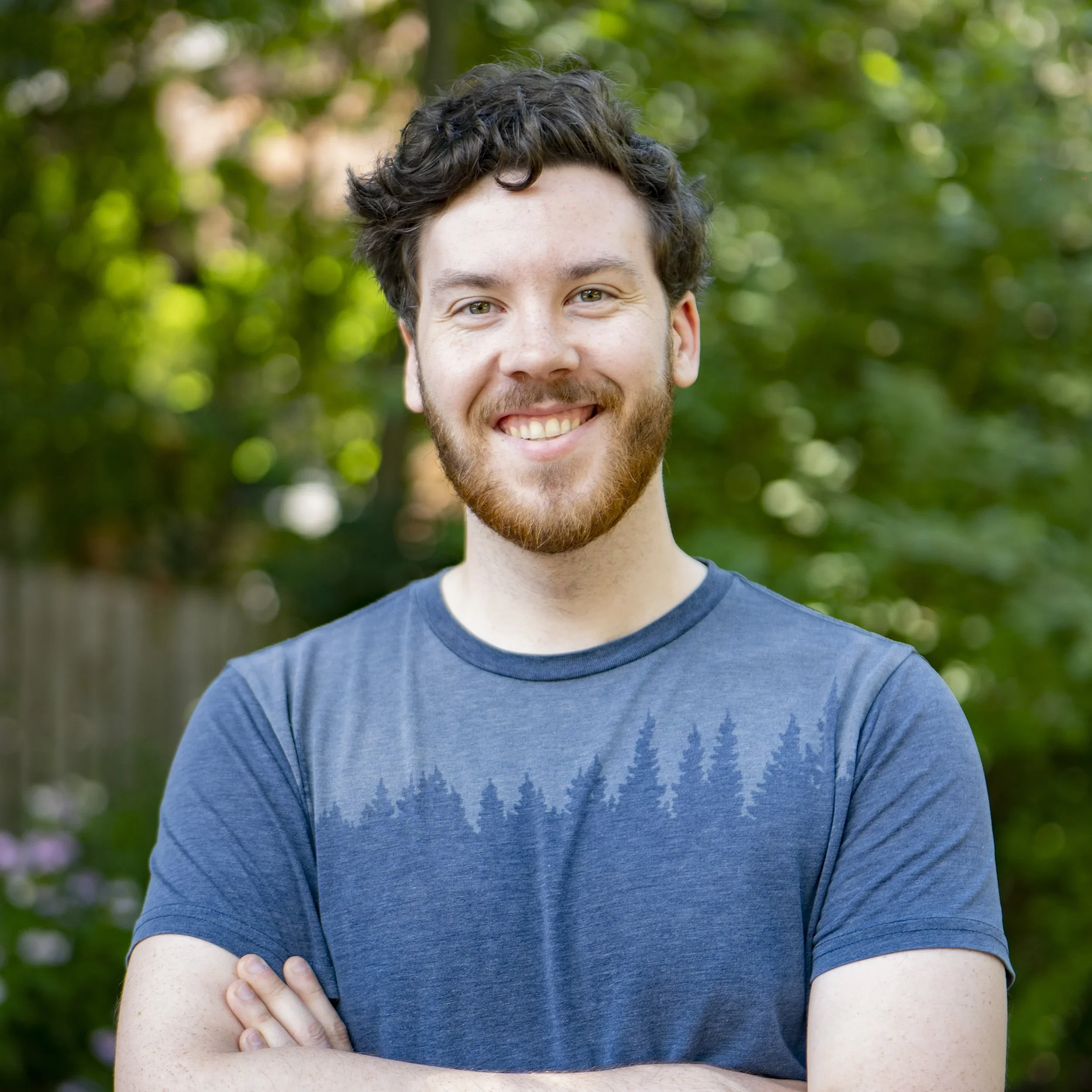 Young man with dark curly hair and a beard smiling outdoors, wearing a blue T-shirt with a pine tree pattern.