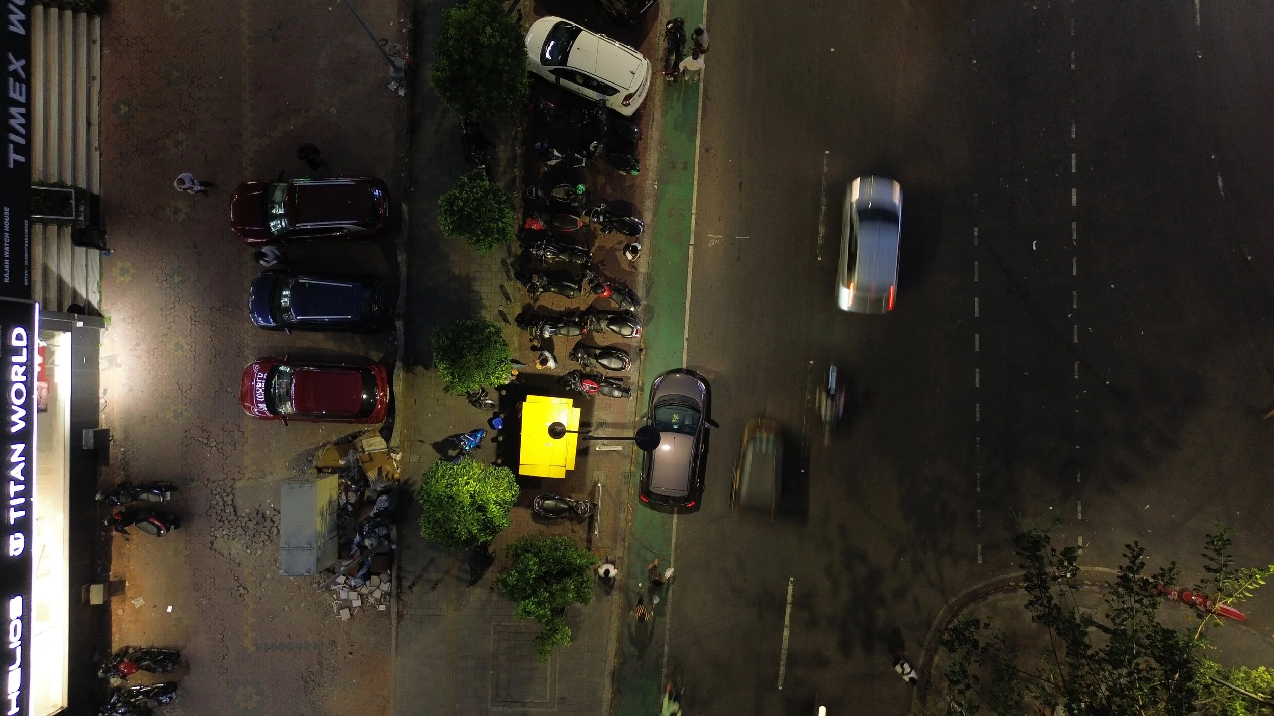 Nighttime view from above of a parking lot and street with moving cars, motorcycles, trees, and a small group of people near parked motorcycles and scooters.