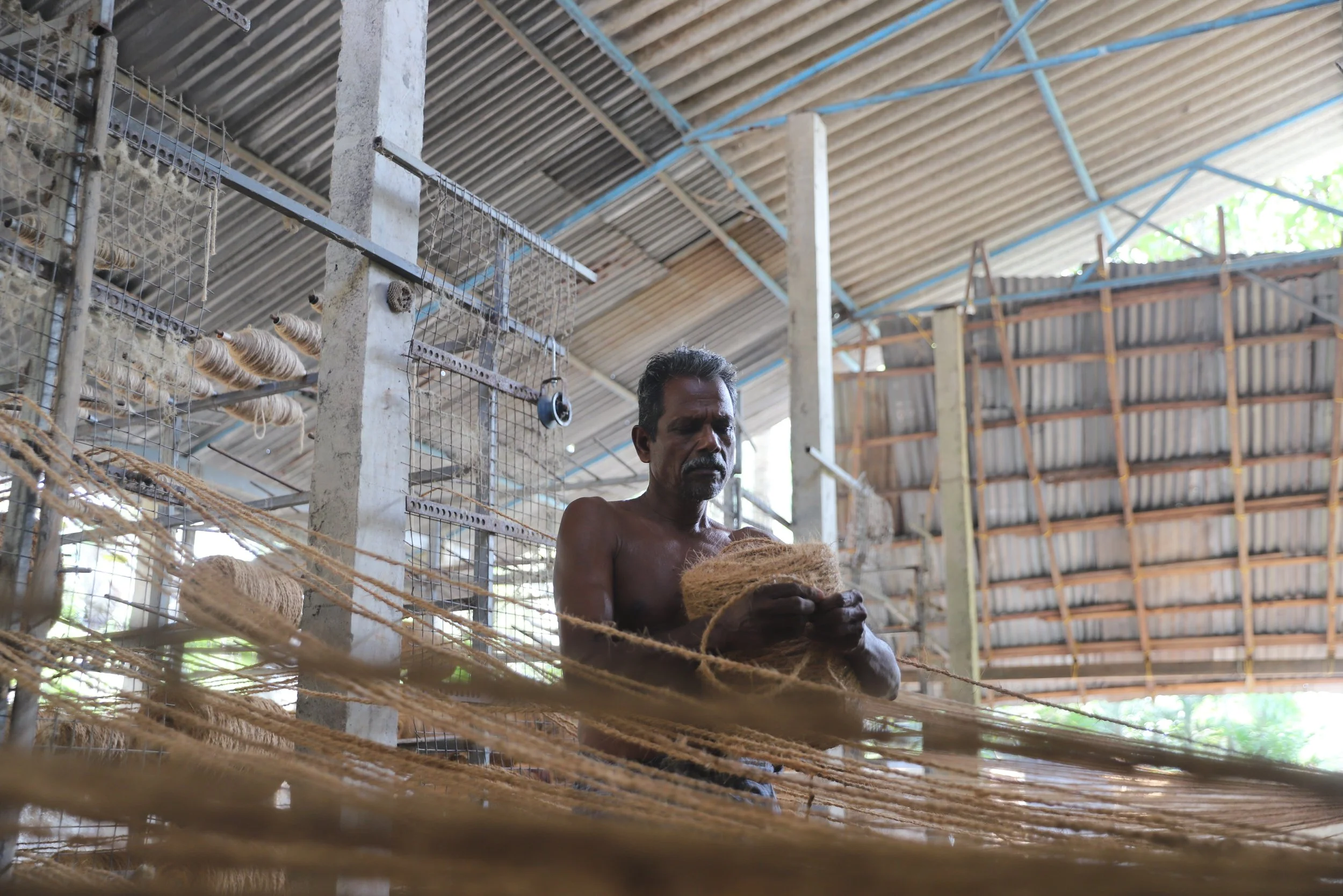 A shirtless man working with fibrous natural material inside a workshop with a metal roof and wire shelving.