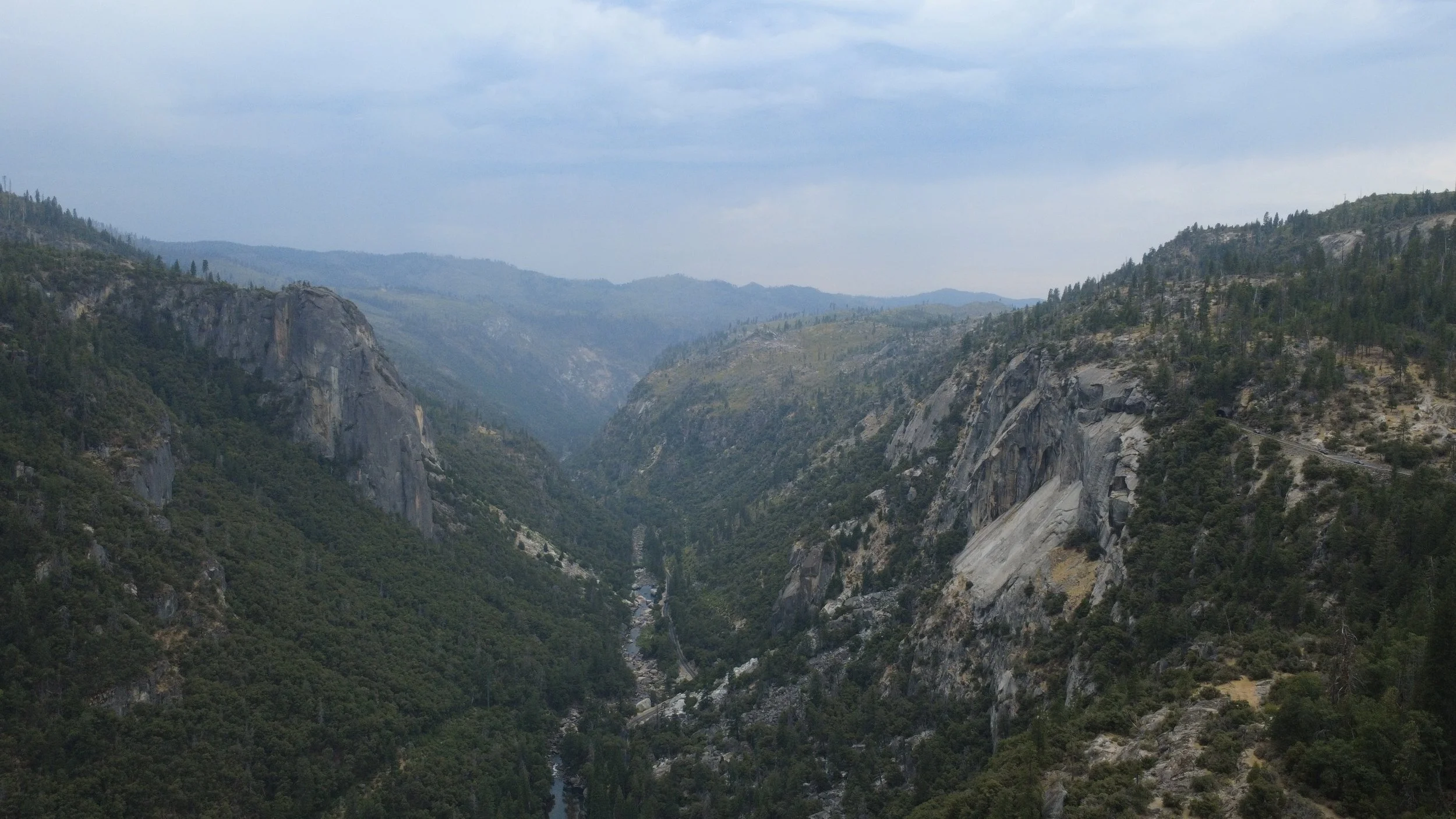 View of a deep canyon with steep rocky cliffs, dense forest, and a river running through the bottom, under a cloudy sky.