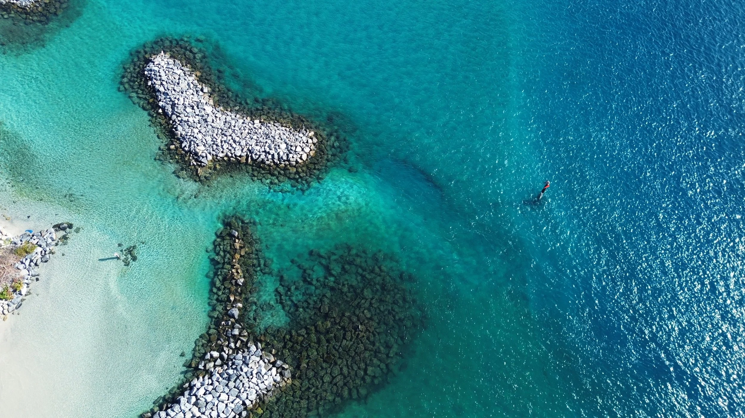 Aerial view of a shoreline with sandy beach, rocky breakwaters, and two paddleboards on turquoise water.