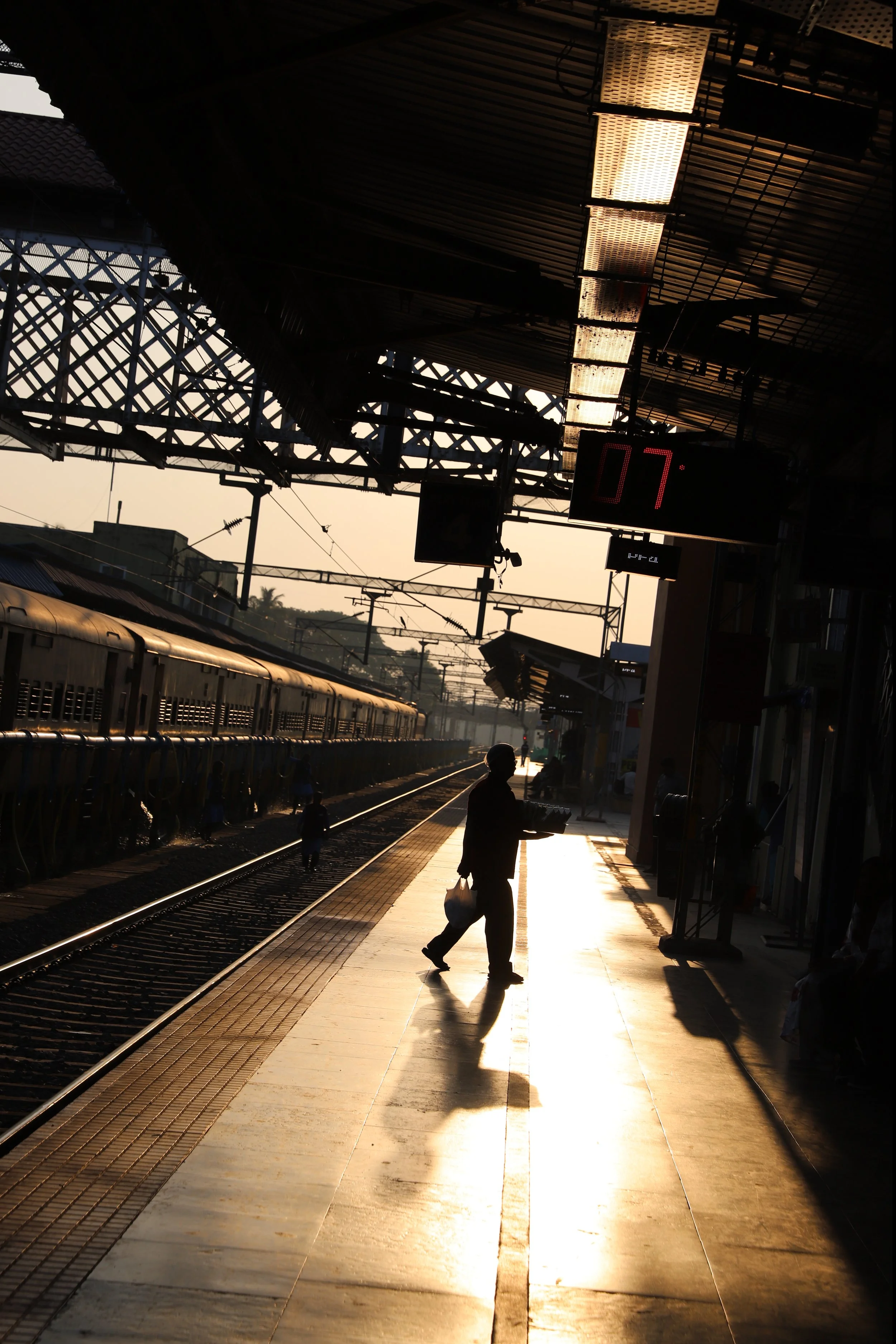 Silhouetted person walking along a train station platform during sunset, with a train on the left and electrical wires overhead.