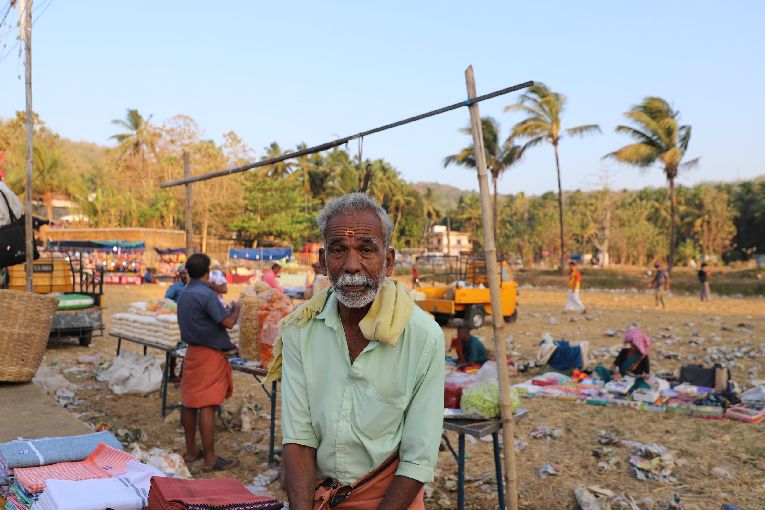 An elderly man with gray hair, a mustache, and traditional attire, standing at an outdoor marketplace with vendors and trees in the background.