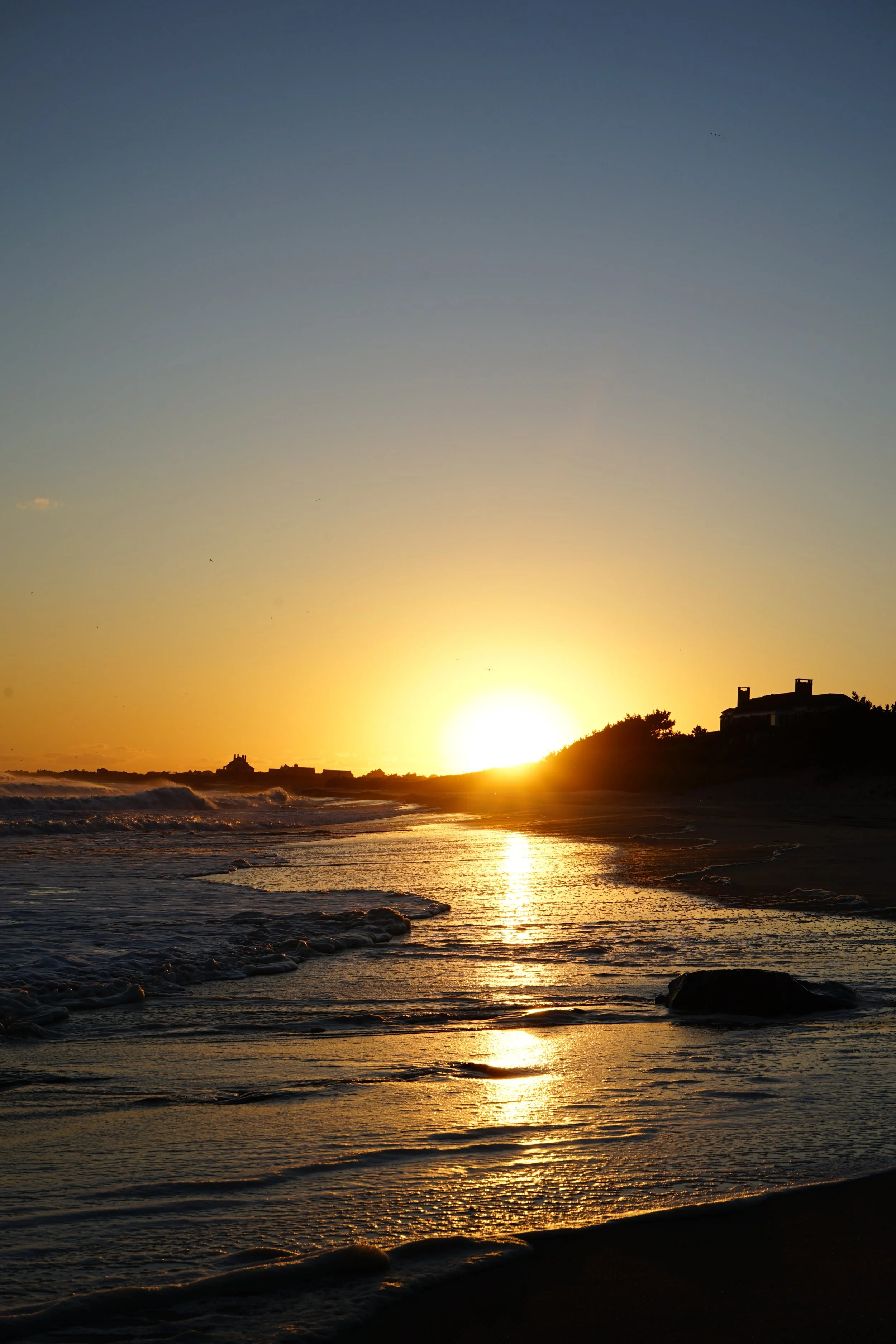 Sunset over the ocean with waves and silhouette of houses on a hill.