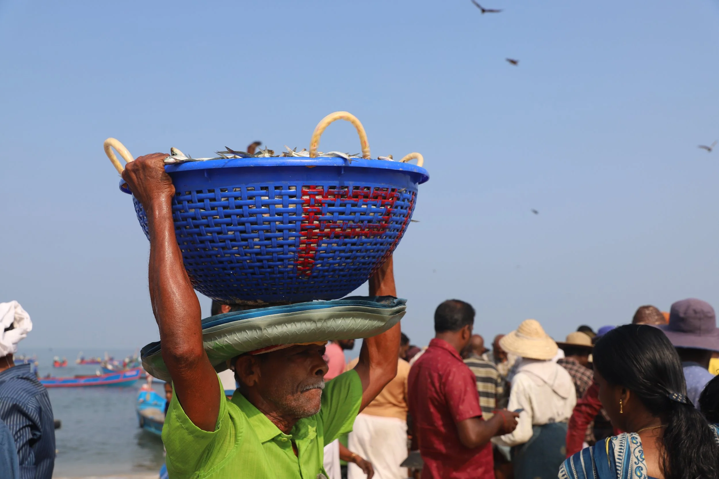 An elderly man with a green shirt balancing a woven basket filled with fish on his head at a crowded fishing harbor, with boats on the water and people in the background.